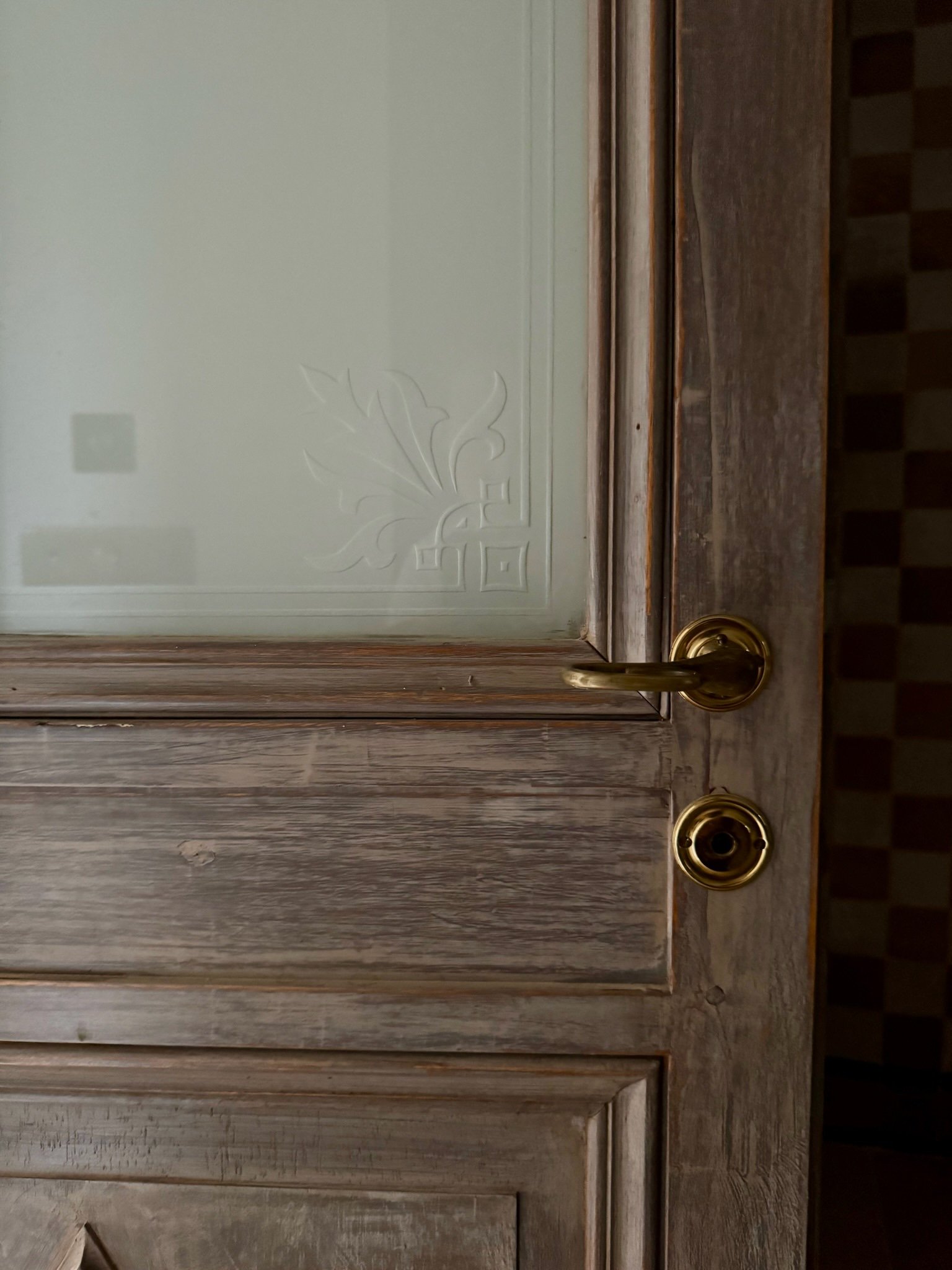A wooden cabinet door with a glass inset, brass handle, and a keyhole, with a patterned tile floor visible in the background.