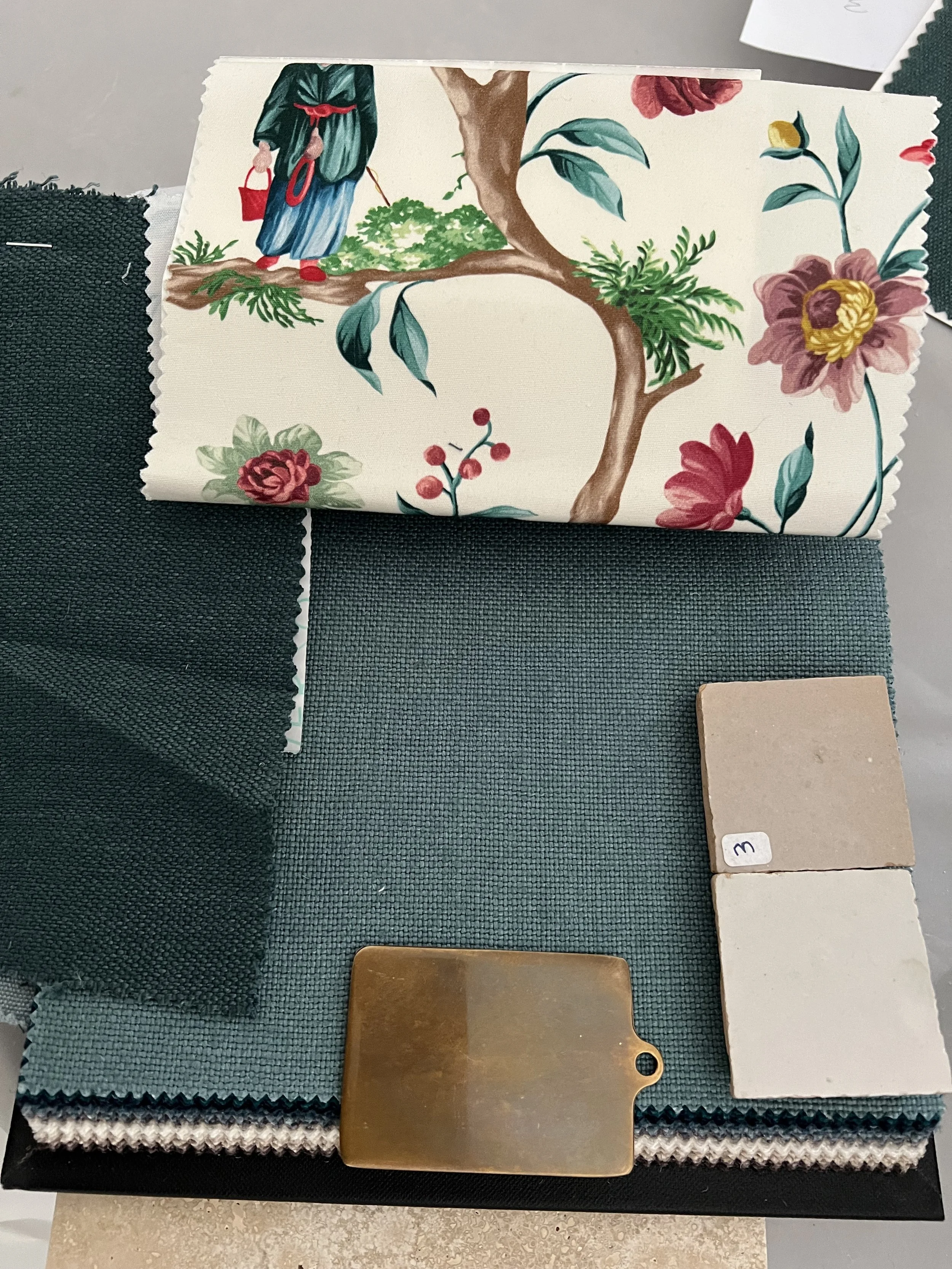 A fabric swatch sample book showing various textile samples, with a floral printed swatch at the top, dark green, teal, and beige fabric samples below, a small beige tile, and a brass clip at the bottom.