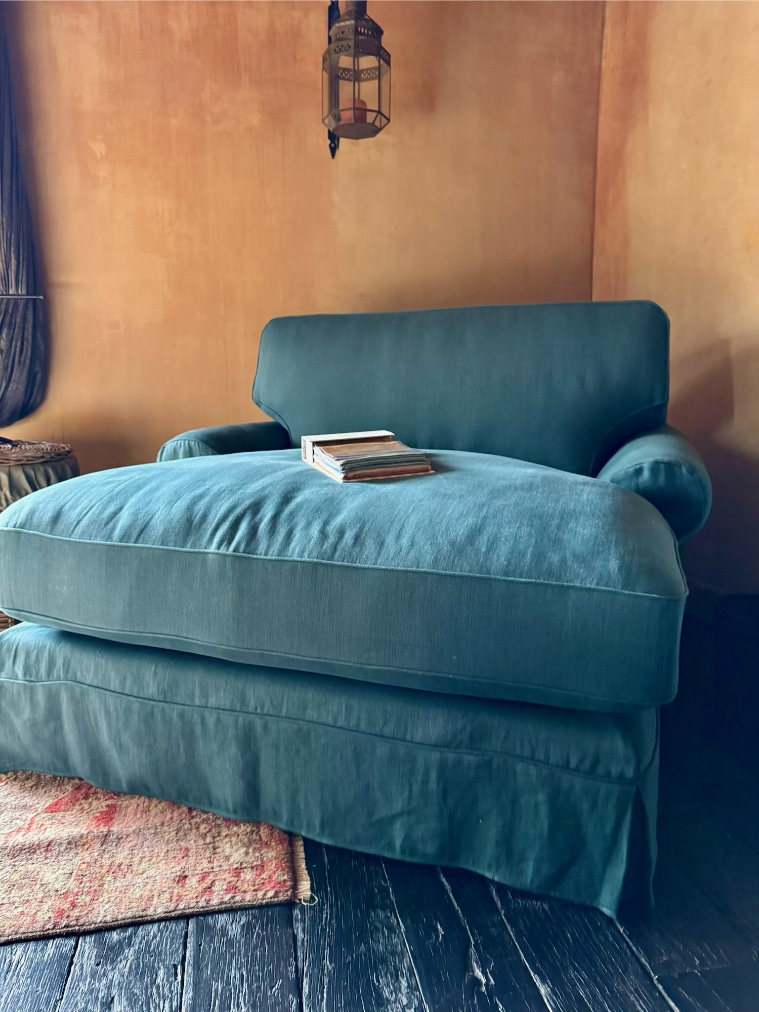 A vintage green velvet sofa with a few magazines on top, set against a brown textured wall with a decorative hanging lantern and a window with dark curtains nearby.