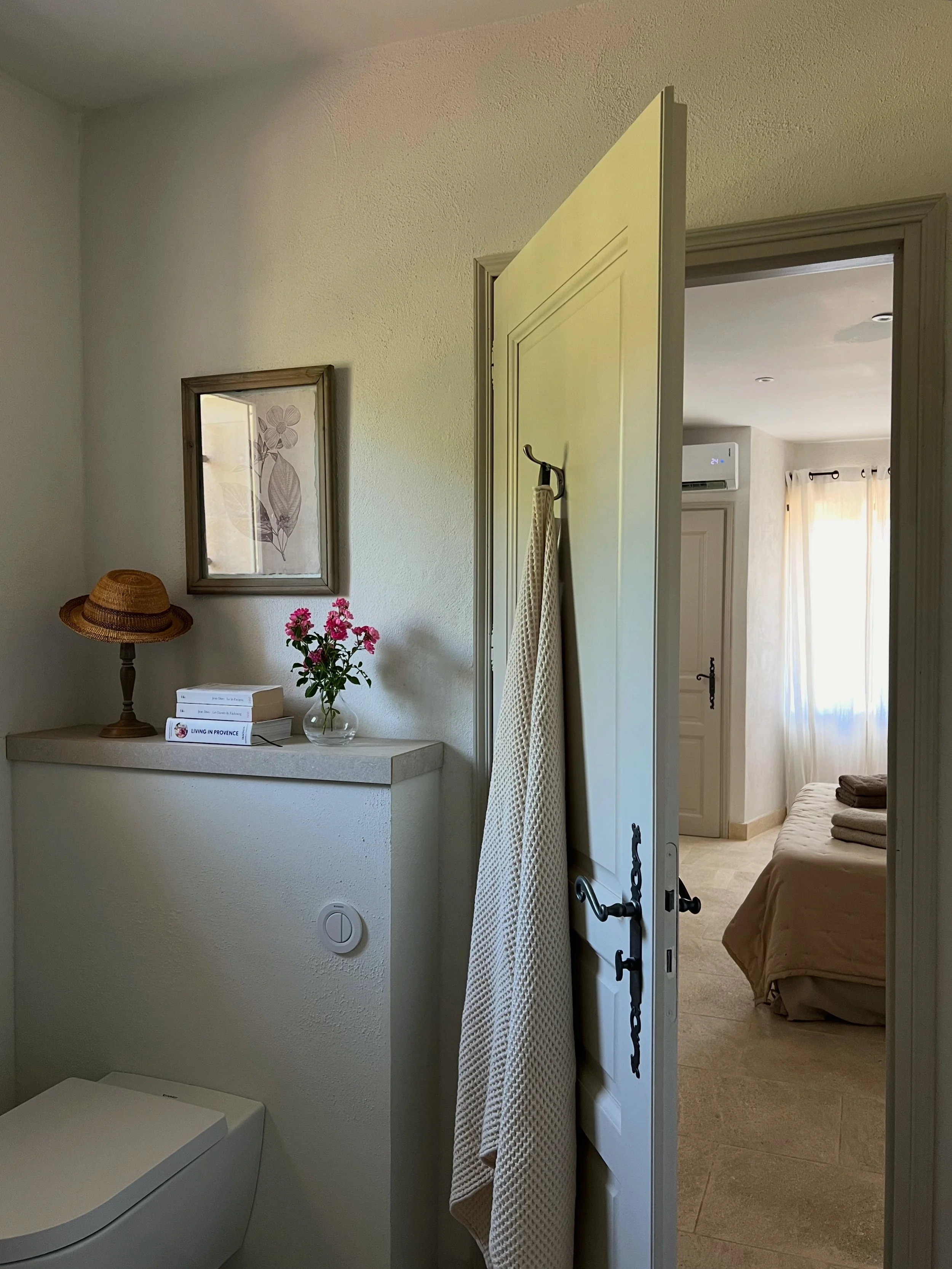 Interior of a small bathroom with a wall-mounted toilet, a shelf with decorative items, including a straw hat, pink flowers in a glass vase, and books; partially open door leading to a bedroom with a bed, folded towels, and sunlight through curtains.