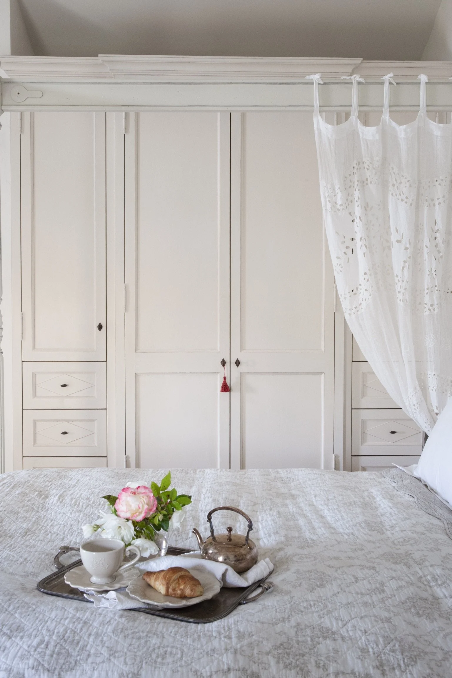 A neatly made bed with a white bedspread, a tray on the bed with a cup, a croissant, a teapot, and a flower arrangement, and a white, wooden wardrobe with decorative drawers and a lace curtain.