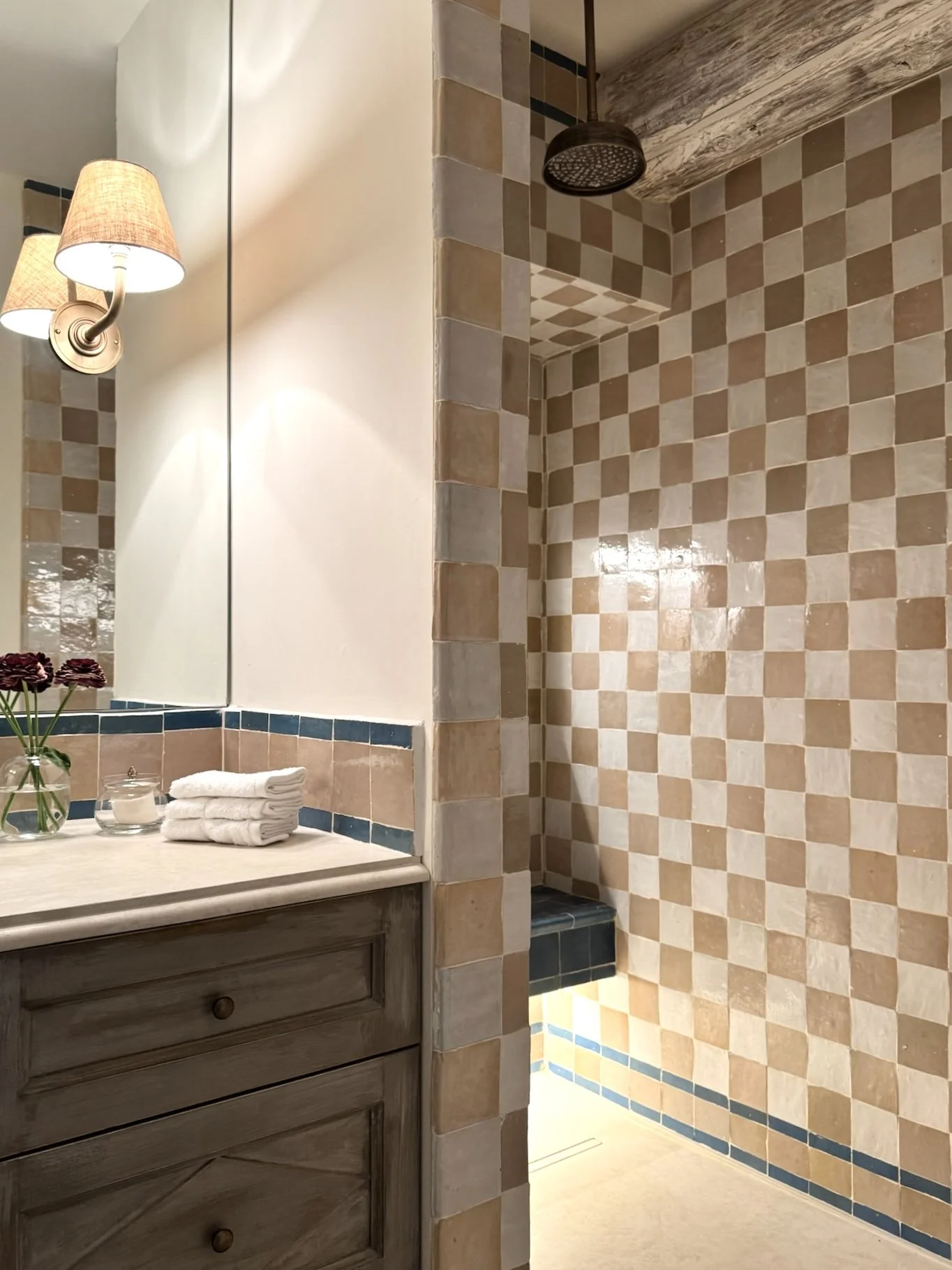A bathroom with a wooden vanity, folded white towels, a small glass candle holder, and a vase with purple flowers on the countertop. The mirror reflects a wall-mounted light fixture with two lampshades. There is a walk-in shower with beige and cream 