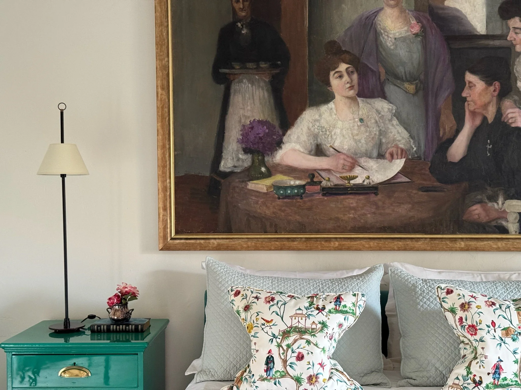 A bedroom with a large framed painting hanging above the bed, showing women and a waiter in a vintage scene. Next to the bed is a turquoise nightstand with a pink flower pot, a book, and a black table lamp. The bed has white pillows with colorful emb