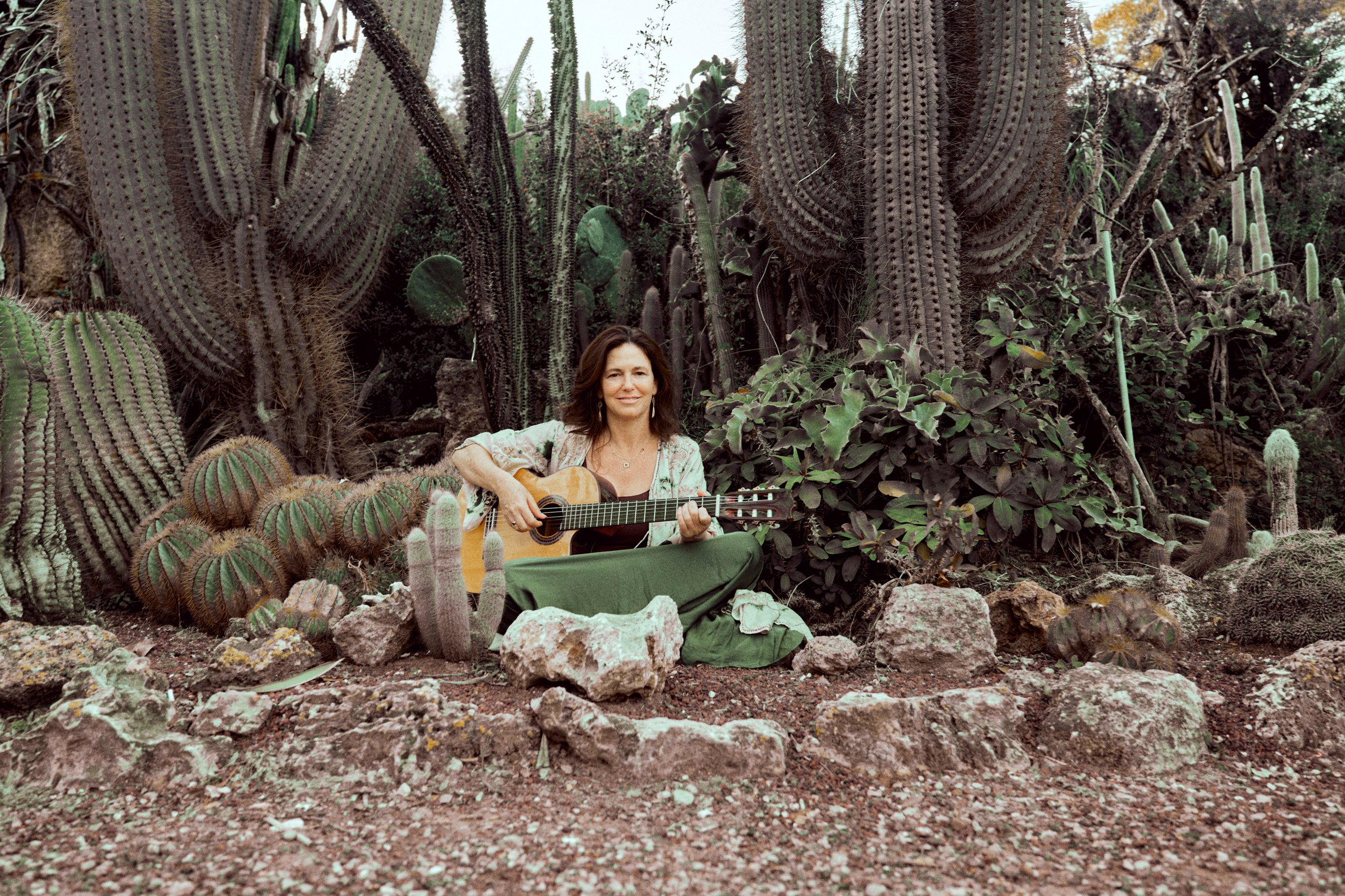 A woman sitting in a cactus garden playing an acoustic guitar.