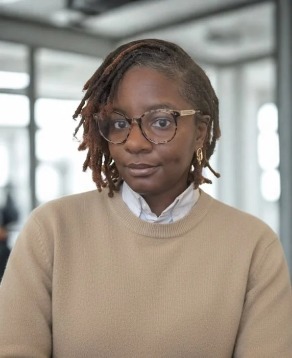 A woman with short brown dreadlocks, wearing glasses, a white collared shirt, and a beige sweater indoors.
