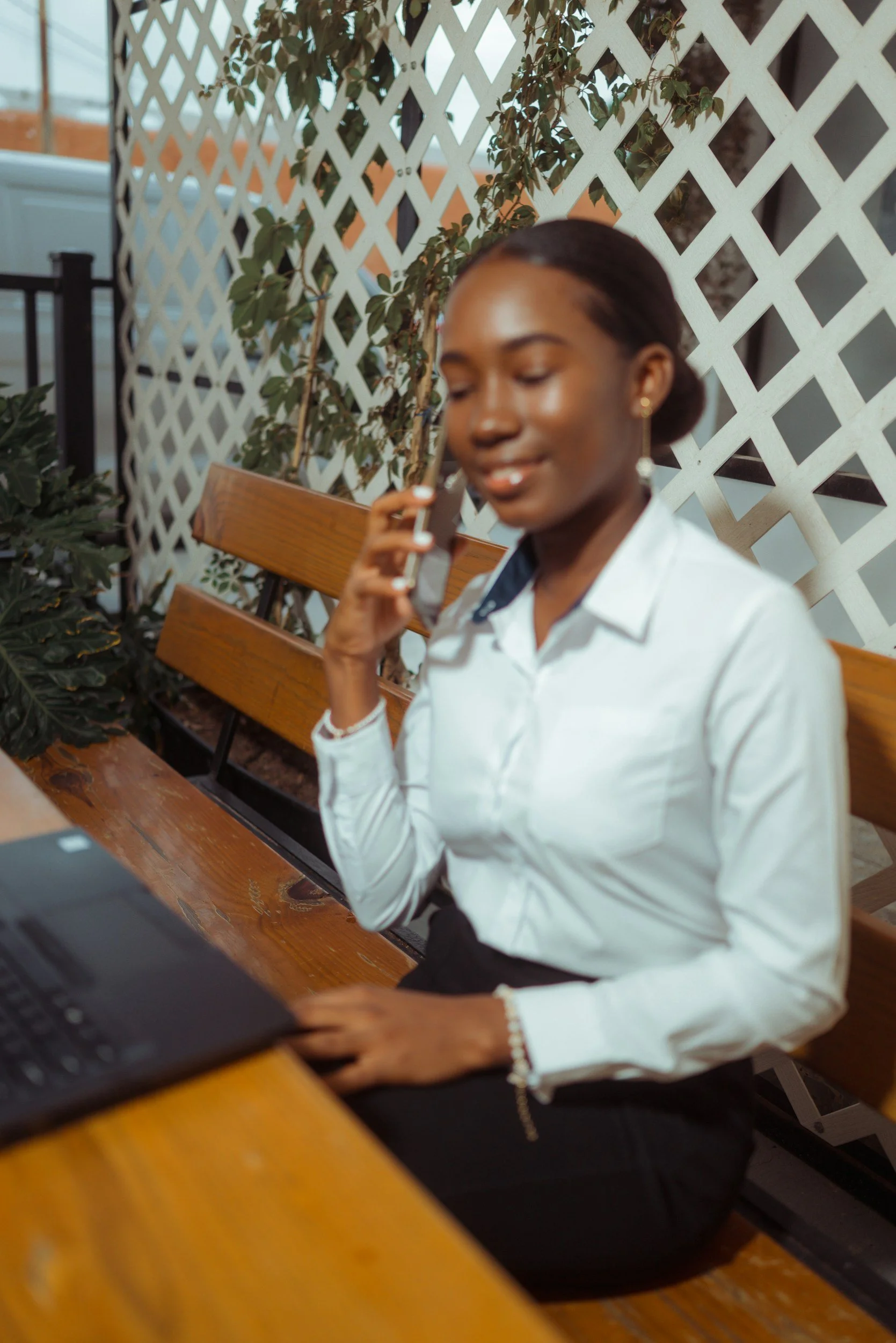 A young woman with dark skin and dark hair tied back, wearing a white blouse and black skirt, sitting on a wooden bench outdoors, talking on her mobile phone while working on a laptop.