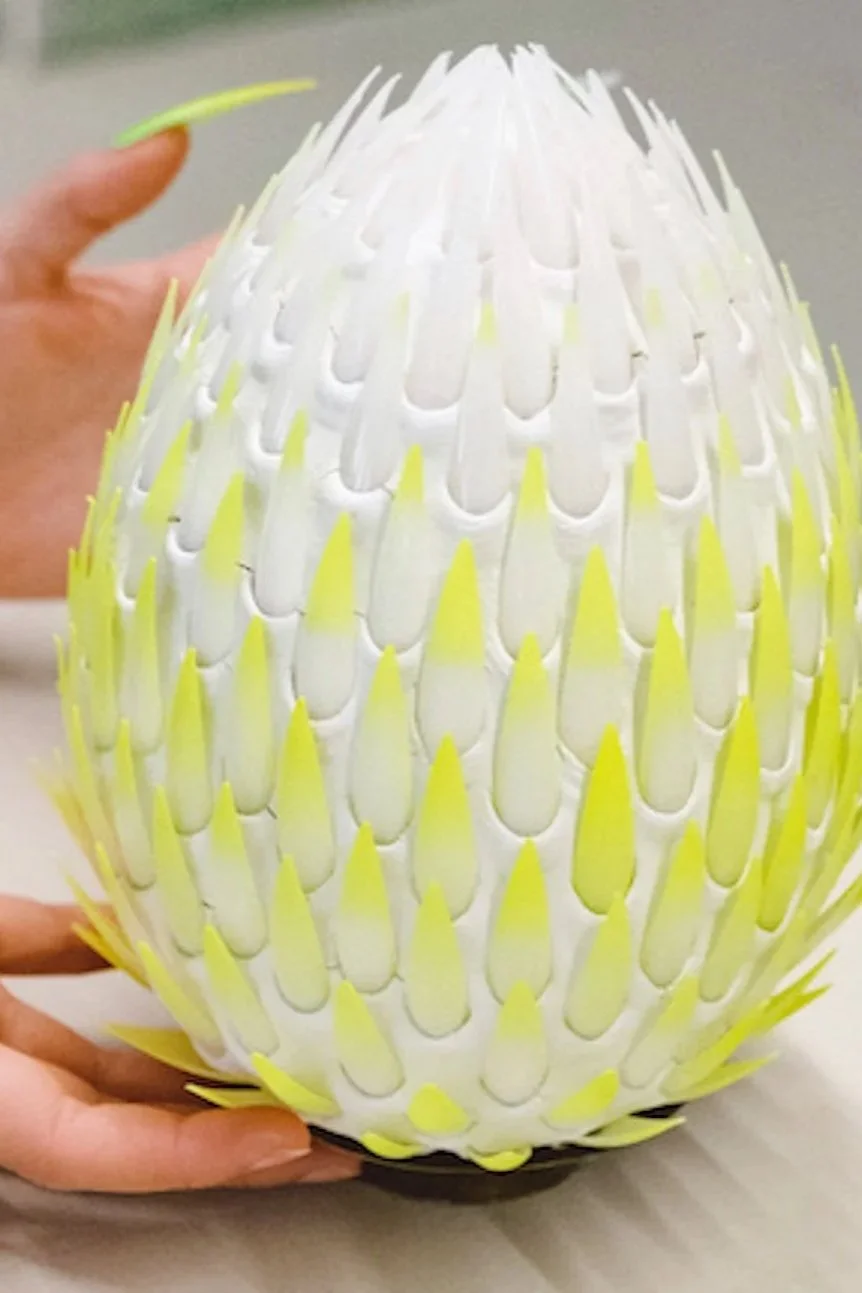 A person holding a decorative egg-shaped object made of white and yellow welded plastic pieces.