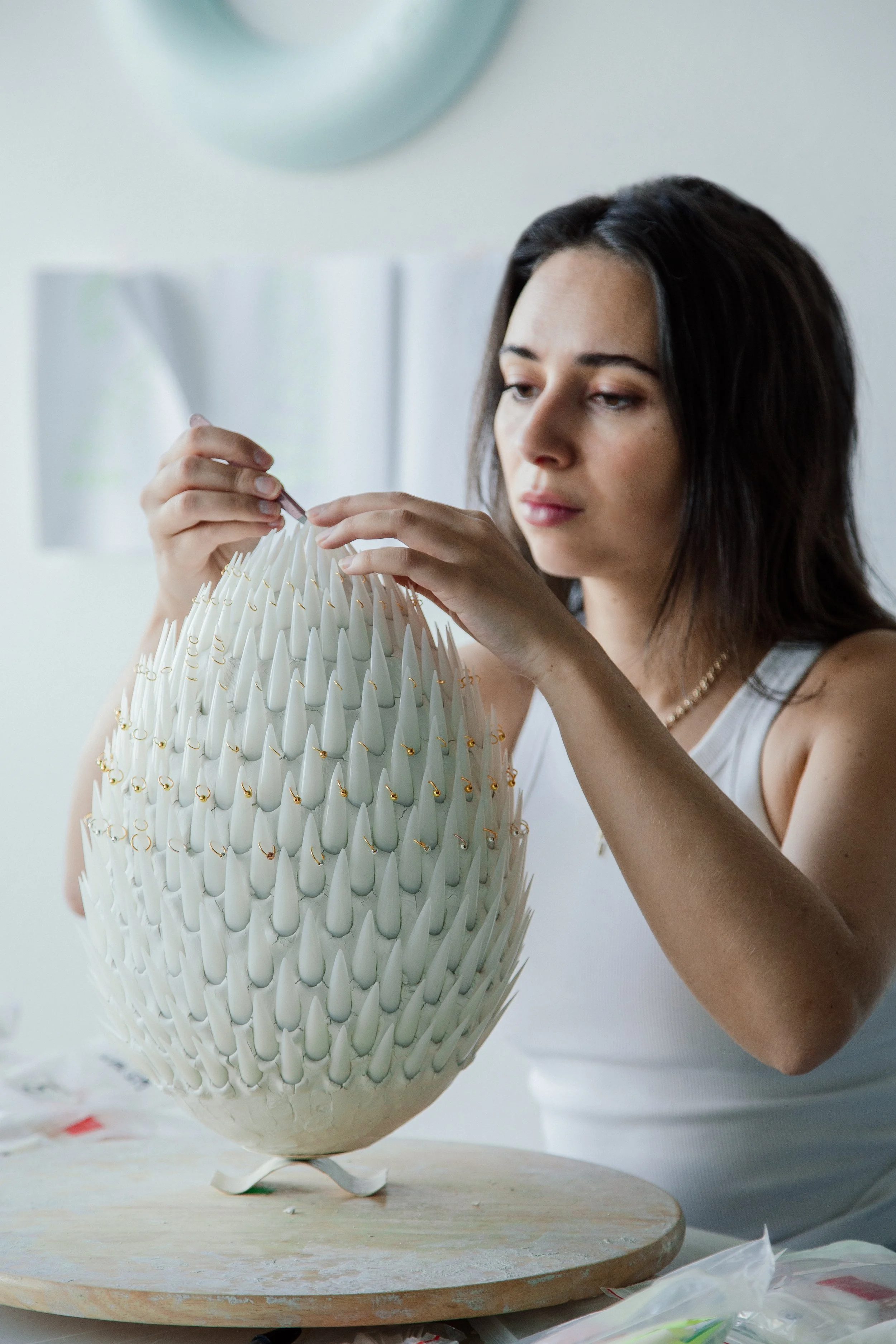 A woman working on a decorative white ceramic or porcelain object with gold accents, possibly a lampshade, in a well-lit room.