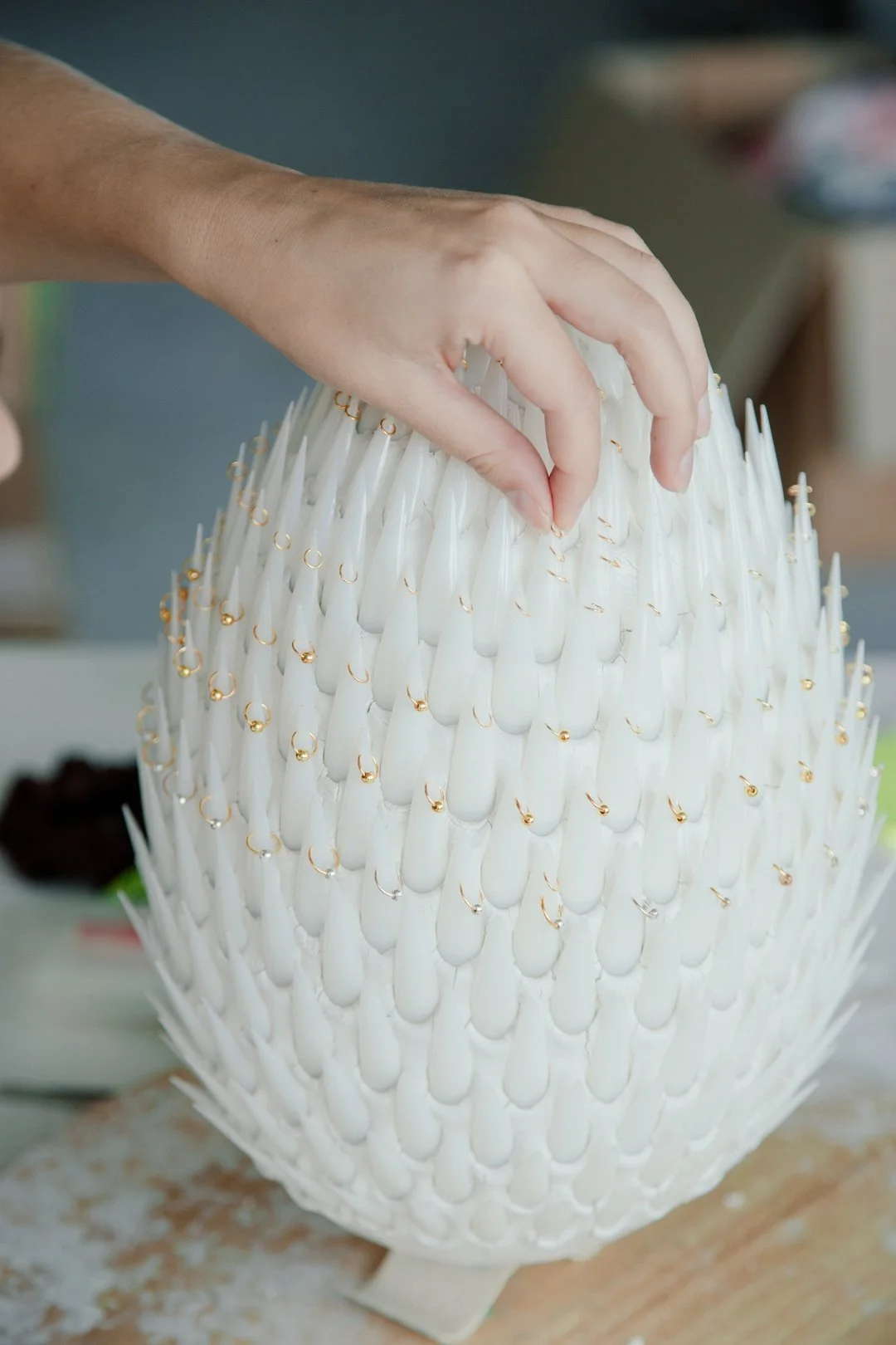 A hand placing a pin into a large white object resembling a pineapple made of many white, petal-like structures with gold accents.