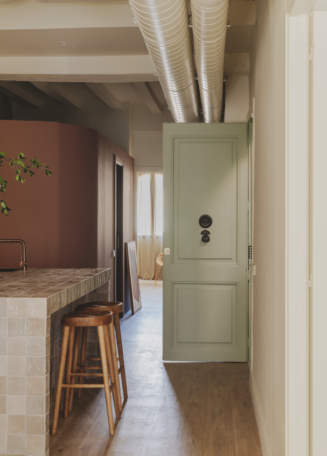 Interior view of a cozy room with a green door, exposed silver ducts, wooden stools at a tiled counter, and a glimpse of a window with curtains.