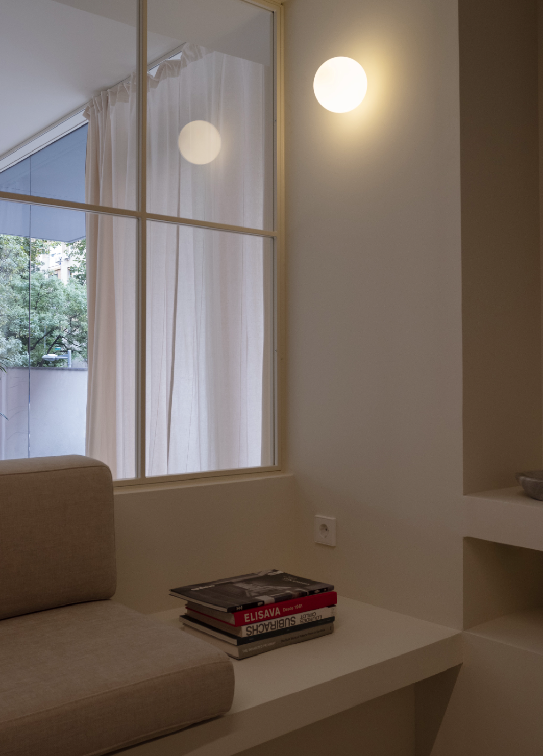 A corner of a room with a large window, a beige cushioned bench, and a stack of books on a built-in shelf. The window has white curtains and a view of trees outside. Two round wall lights are on, illuminating the wall.