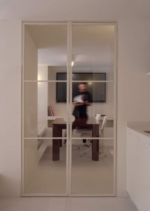 A glass sliding door with white framing, reflecting a person standing inside a room with a dining table and chairs, a TV mounted on the wall, and a bookshelf.