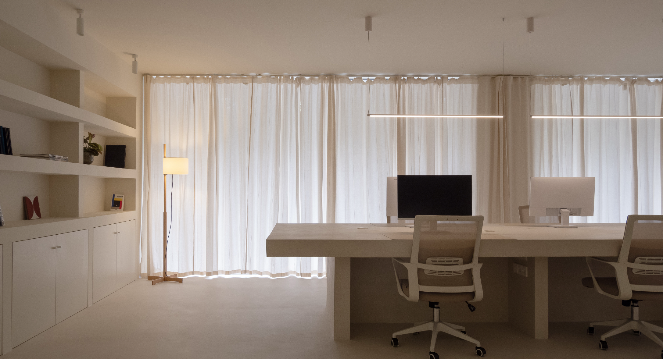 A modern office with large window curtains, two white desks with monitors, beige chairs, a floor lamp, and wall shelves with books and decorative items.