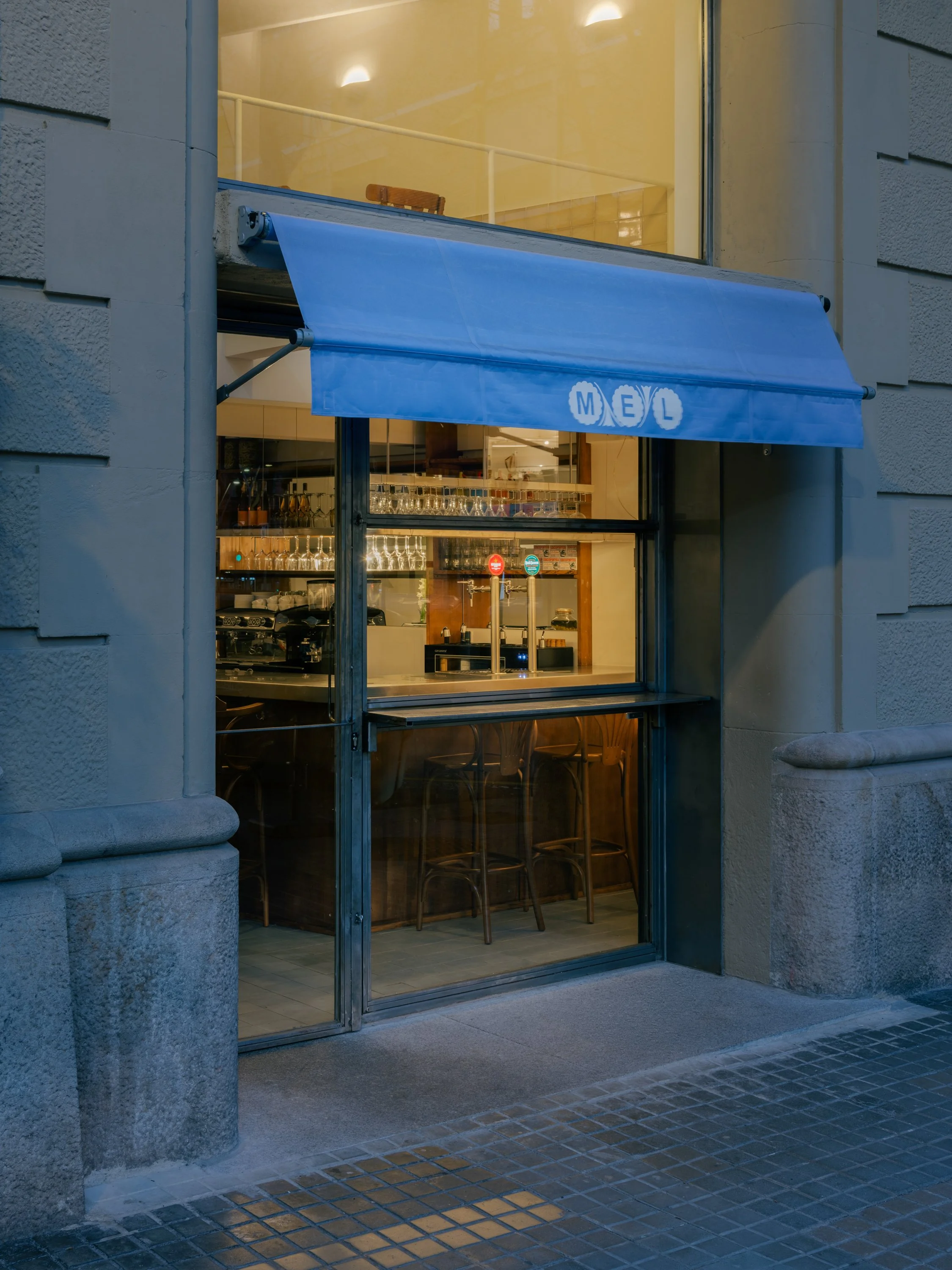 Front view of a closed storefront with a large glass window and a blue retractable awning with the word 'MEL' on it, on a city sidewalk with tiled paving.