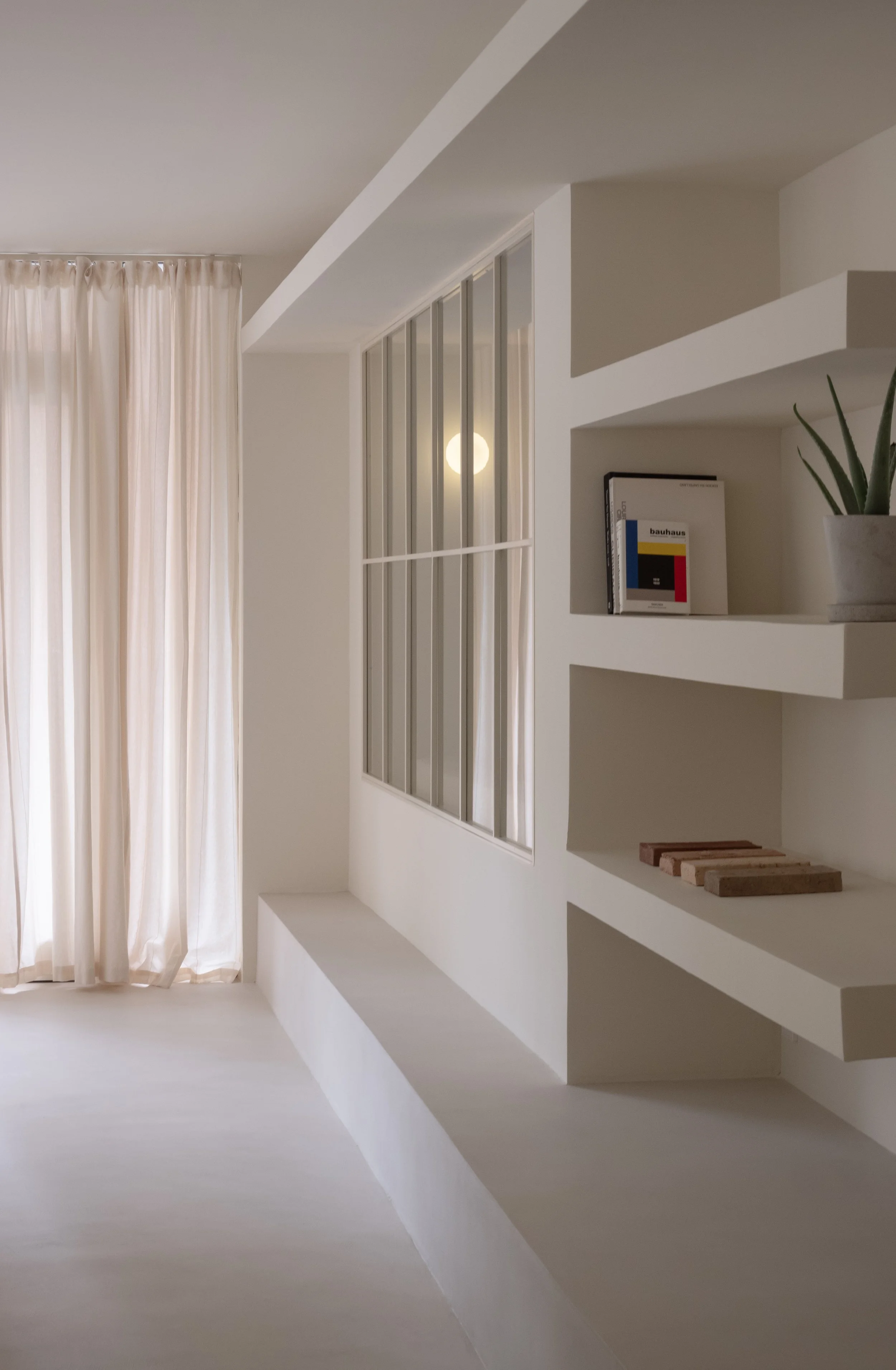 A minimalist interior with beige curtains on the left, white walls, a window with vertical blinds, built-in shelving with books, a potted plant, and some wooden samples on the right.