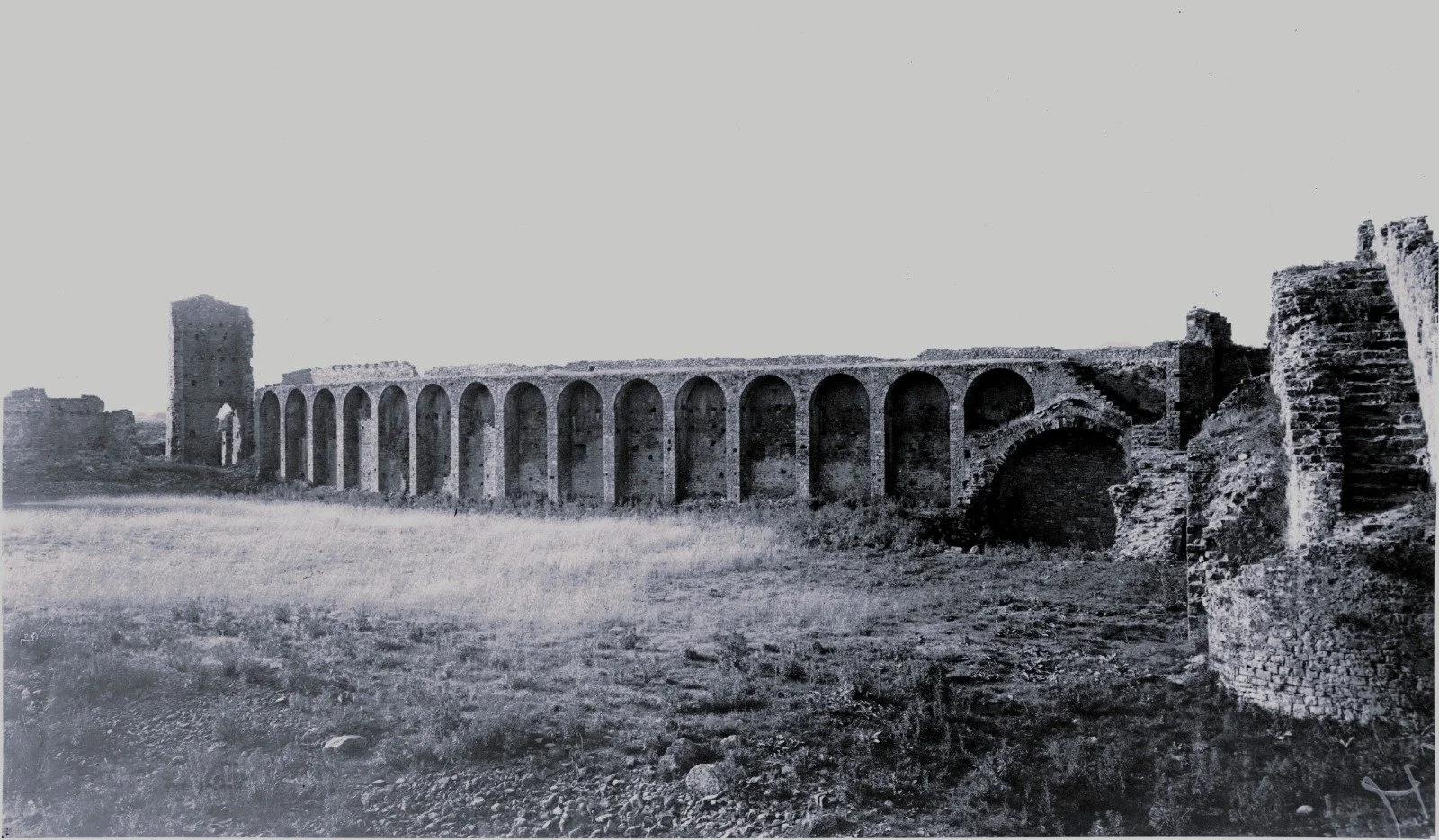 Black and white photo of ancient stone aqueduct with multiple arches, ruins on the right, and open grassy area in the foreground.
