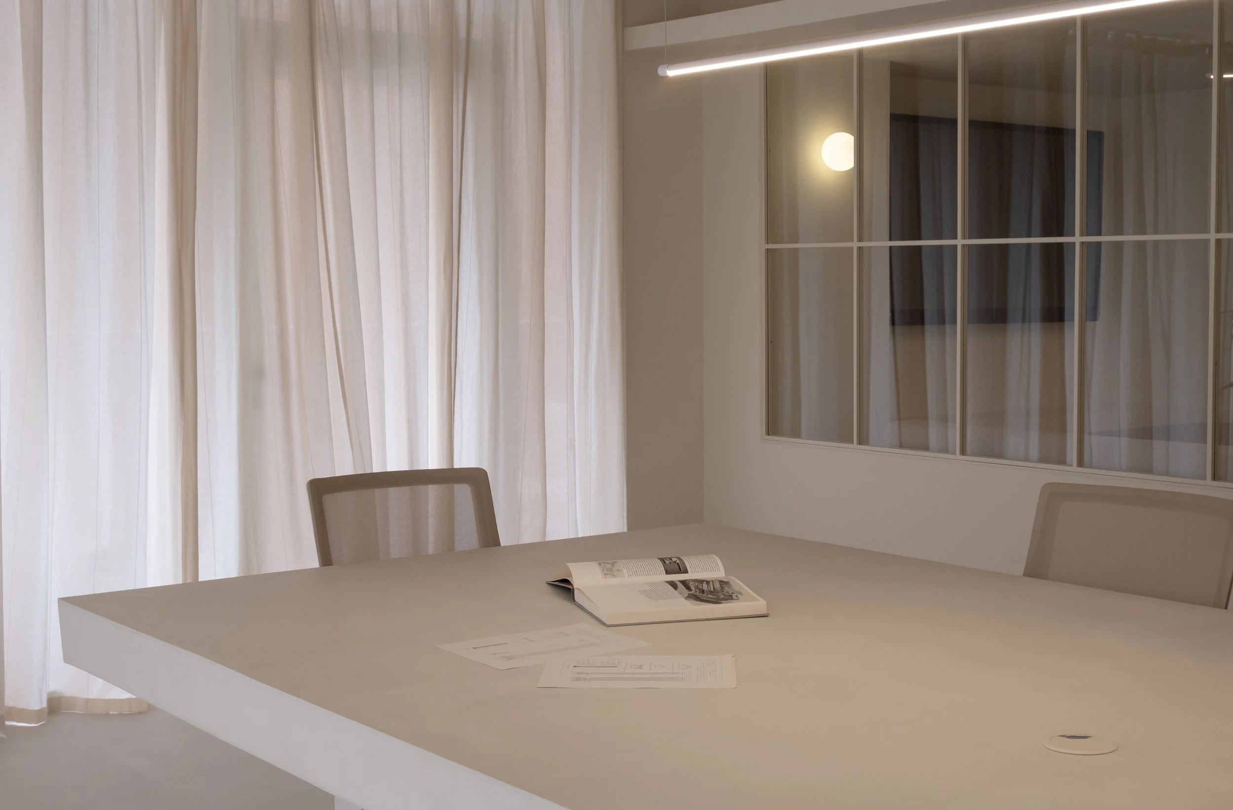 Modern conference room with a white table, beige chairs, open magazine, large window with sheer curtains, and glass wall with a reflection of a round wall lamp.