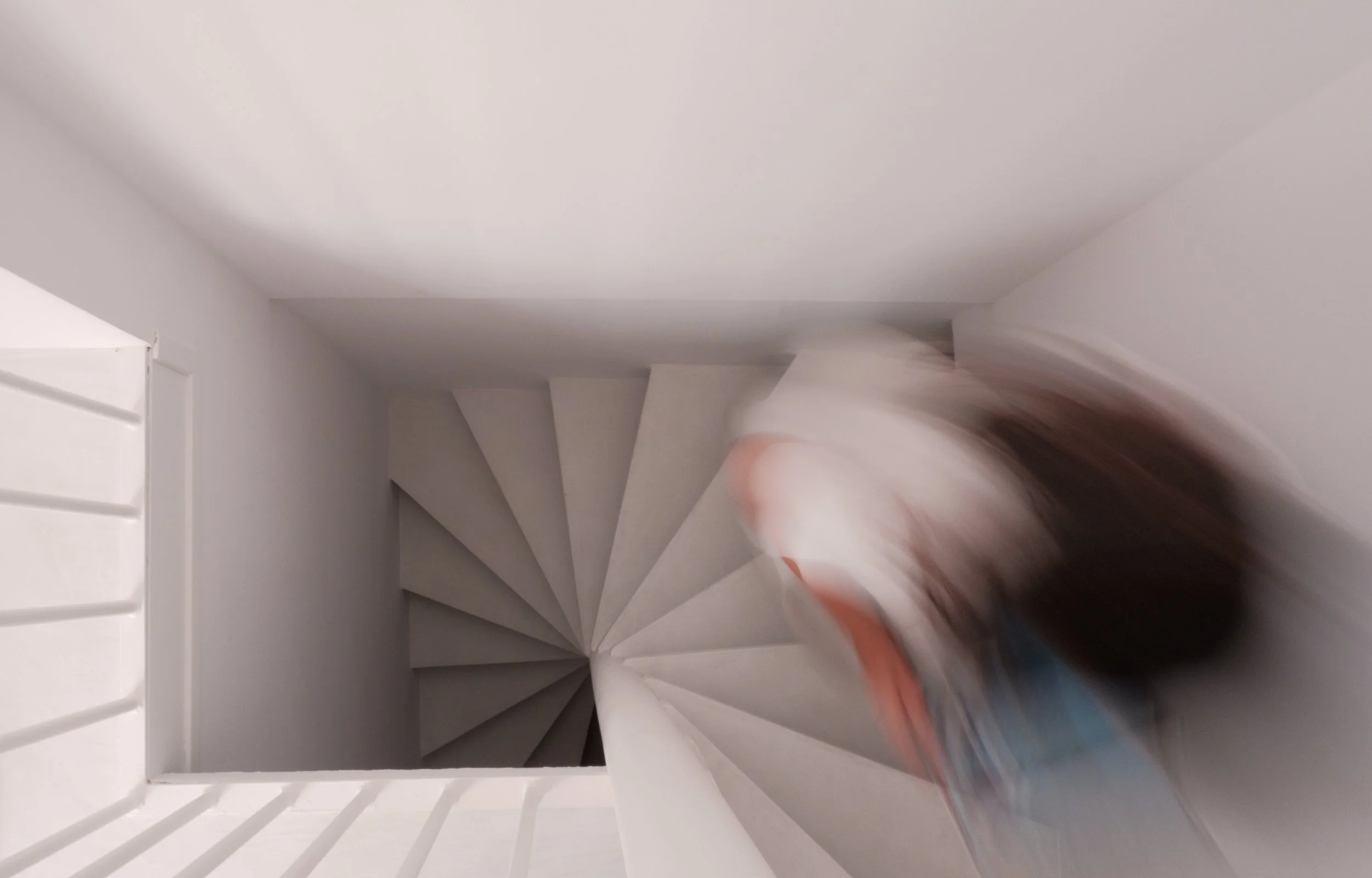 A spiral staircase viewed from above, with white walls and a person climbing the stairs, wearing blue jeans and a white shirt.