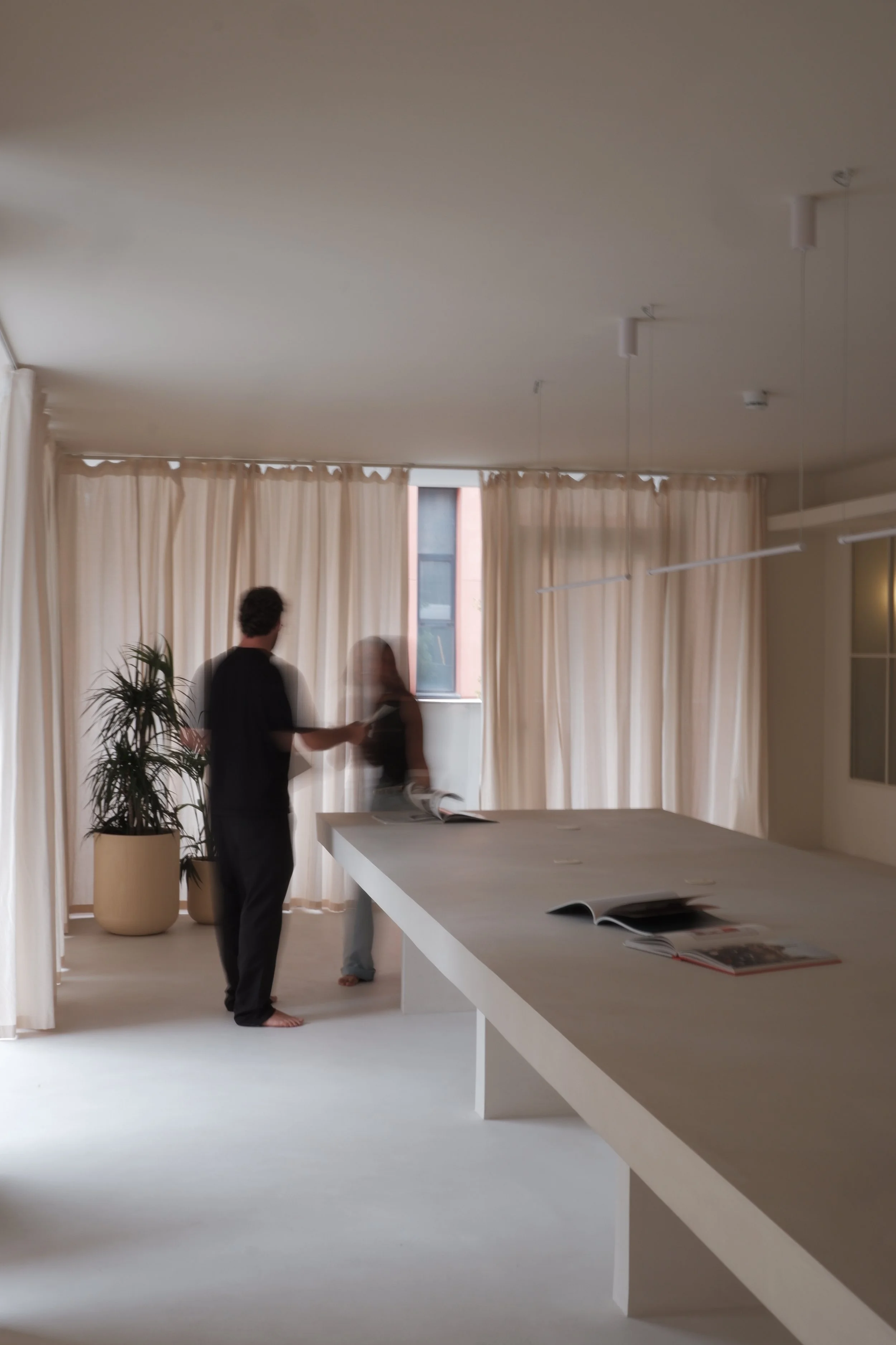 Three people in a well-lit room with large windows and beige curtains, engaging in conversation near a white table with open magazines, a tall potted plant nearby, and ceiling lighting.