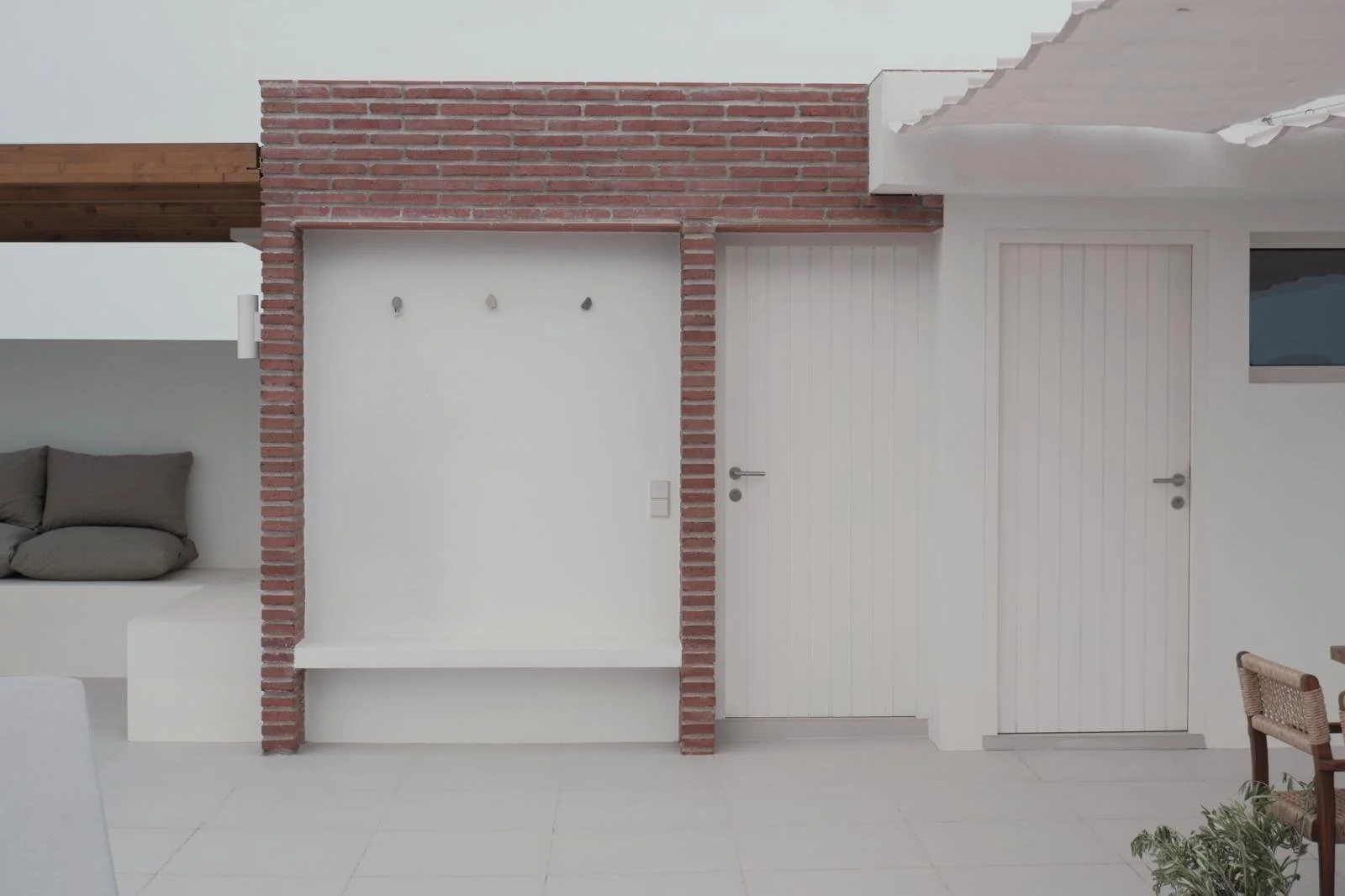 Interior view of a modern room with white walls, a brick column, two white doors, a small seating area with gray cushions, and a window with a view outside.