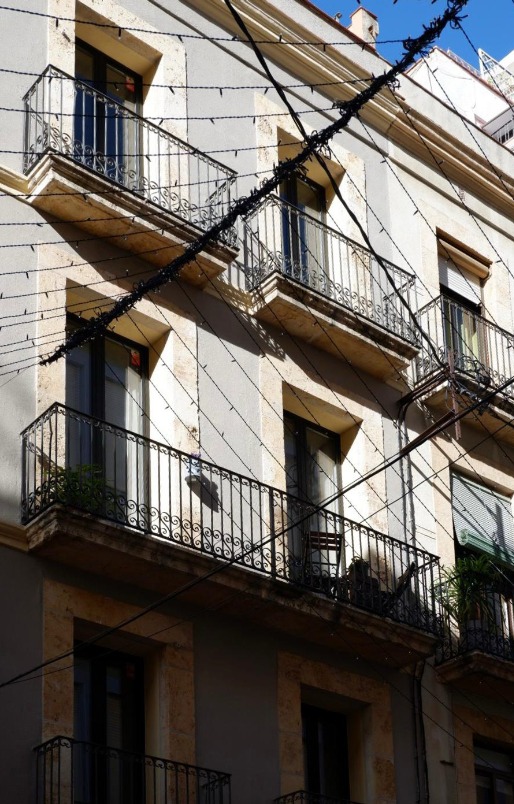 Exterior view of an apartment building with multiple balconies, some with potted plants, electrical wires crisscrossing in front, under a clear blue sky.