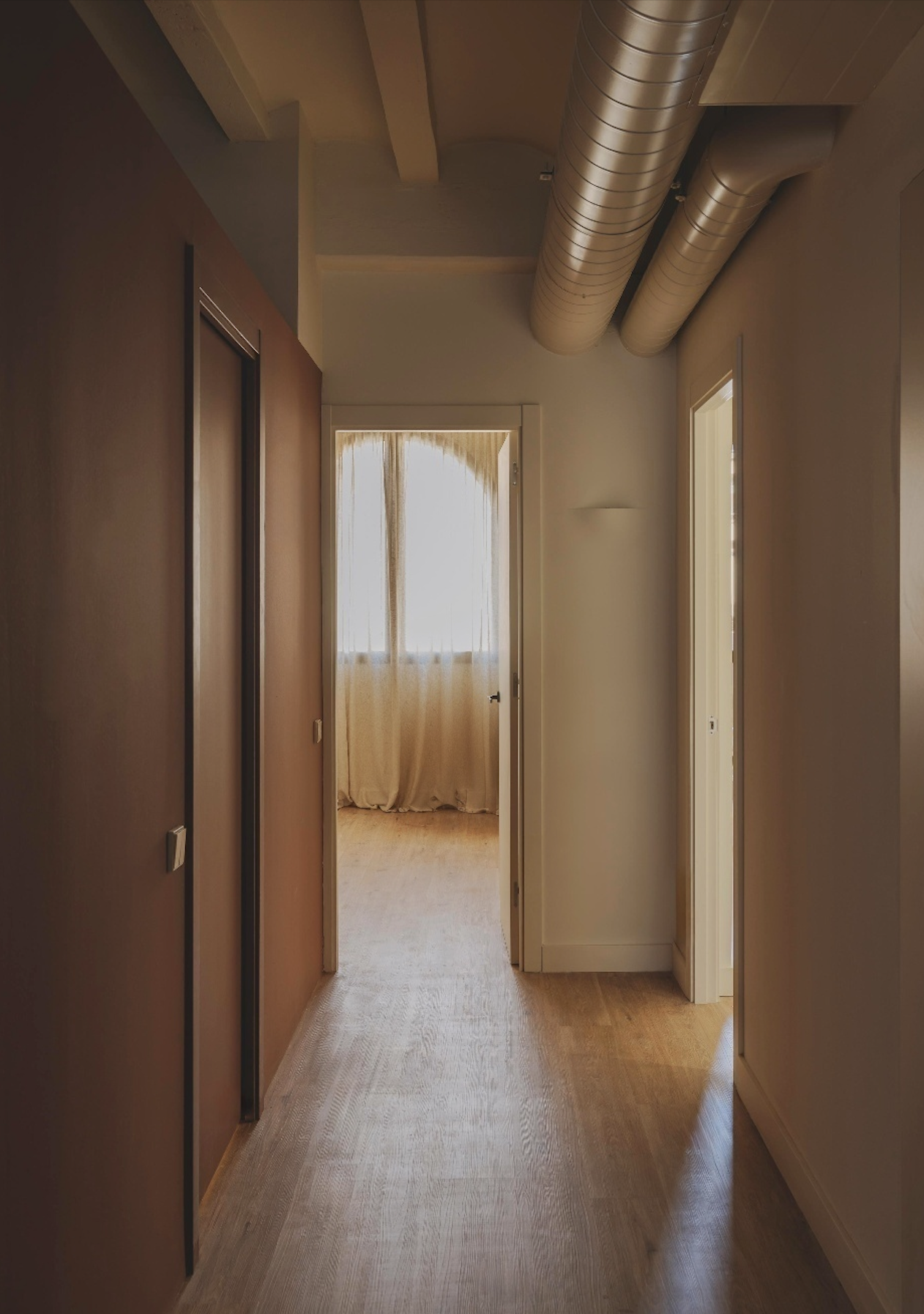 A hallway with wooden flooring, white and brown walls, and a white door at the end with a curtain covering a window. Air conditioning ductwork is visible on the ceiling.