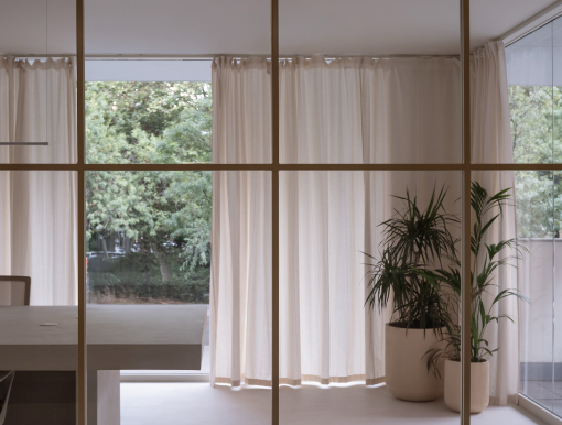 Interior view of a room with large windows, white curtains, and potted plants, overlooking green trees outside.