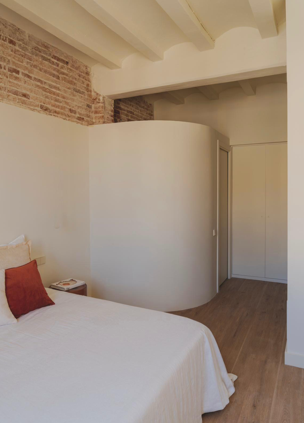 A bedroom with a white bed, a red pillow, a bedside table with a book, an exposed brick wall, a curved white wall, and a cream-colored wardrobe.