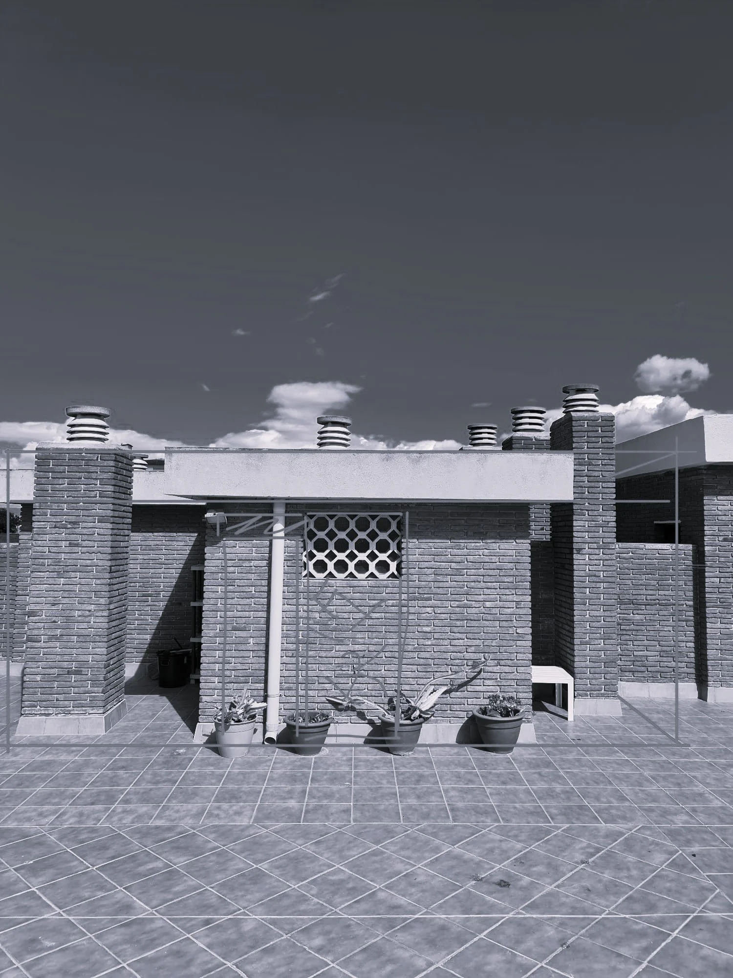 A rooftop terrace with brick columns, potted plants, and a tiled floor, under a clear sky with scattered clouds.