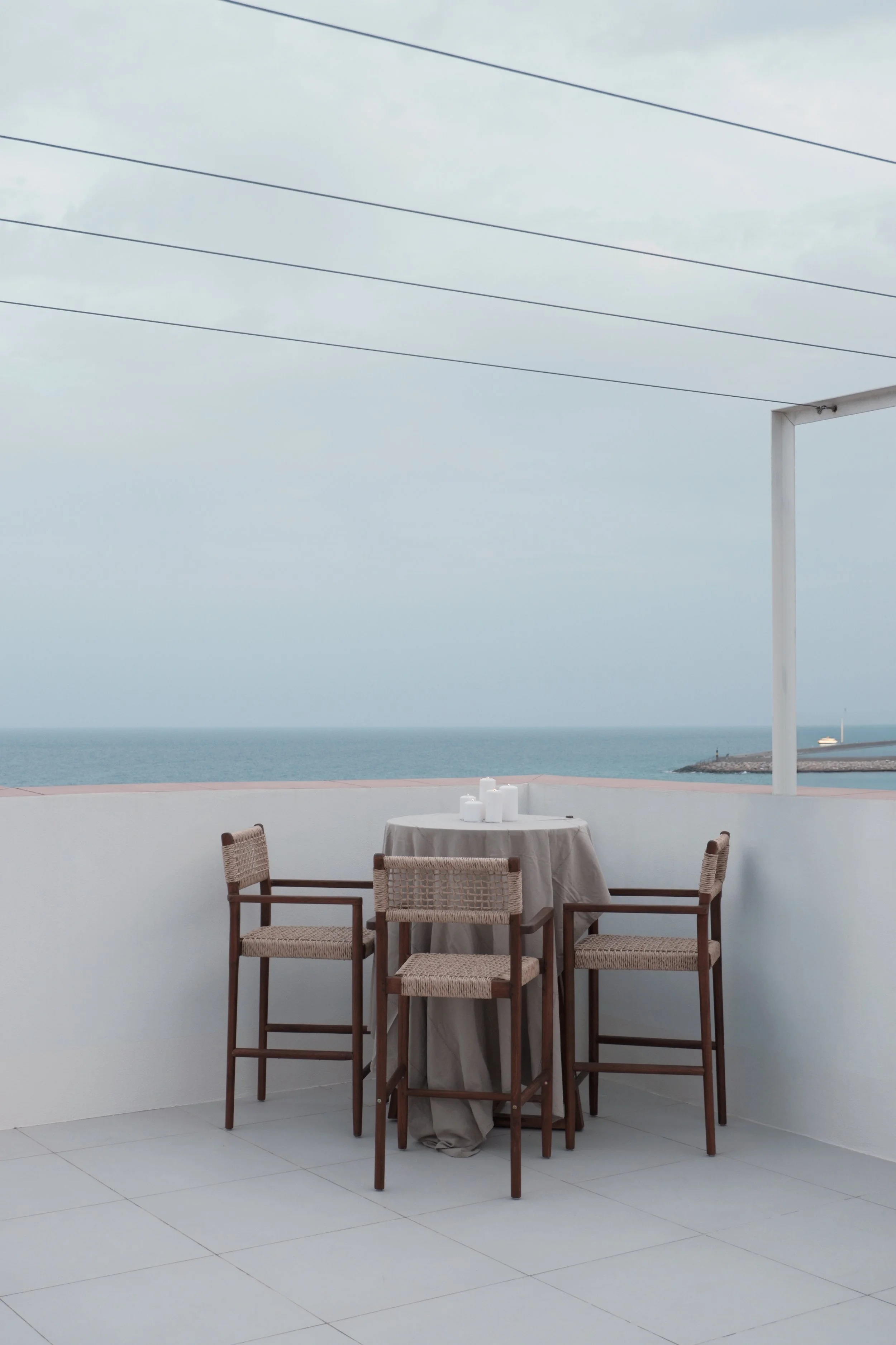 Outdoor dining area with a round table draped in a beige cloth, surrounded by four wicker chairs with wooden frames, overlooking the ocean with a cloudy sky.