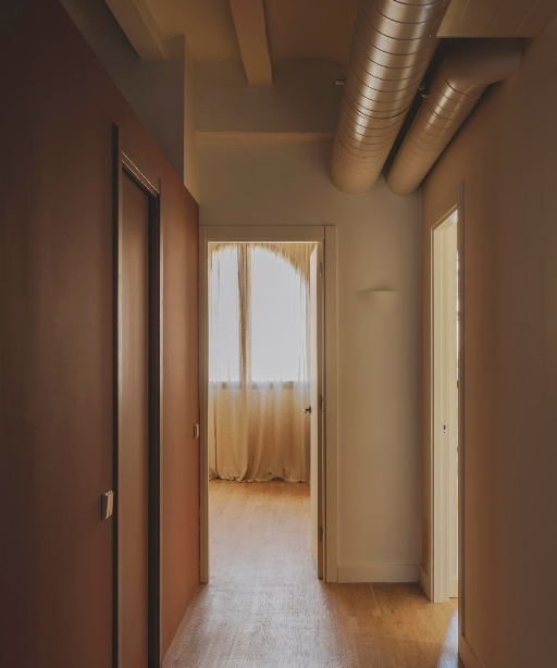 View of a hallway leading to a sunlit room with curtains, exposed ductwork on the ceiling, and wooden flooring.