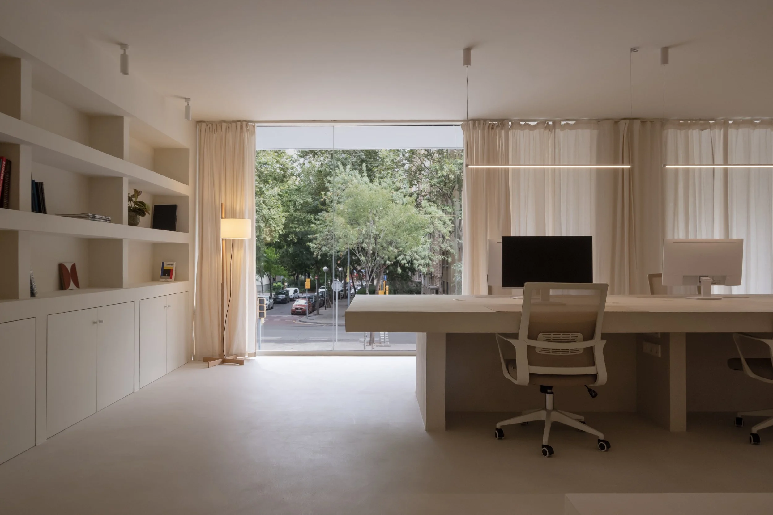 Modern office space with large window showing trees outside, white desk with white chair, computer, and minimalistic decor.