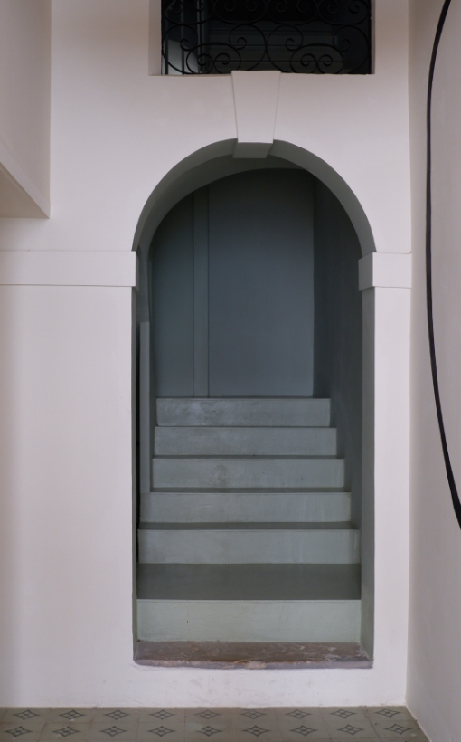 A narrow arched doorway leading to a stairway with six steps, painted in shades of blue, with a small balcony and decorative iron railing visible at the top.