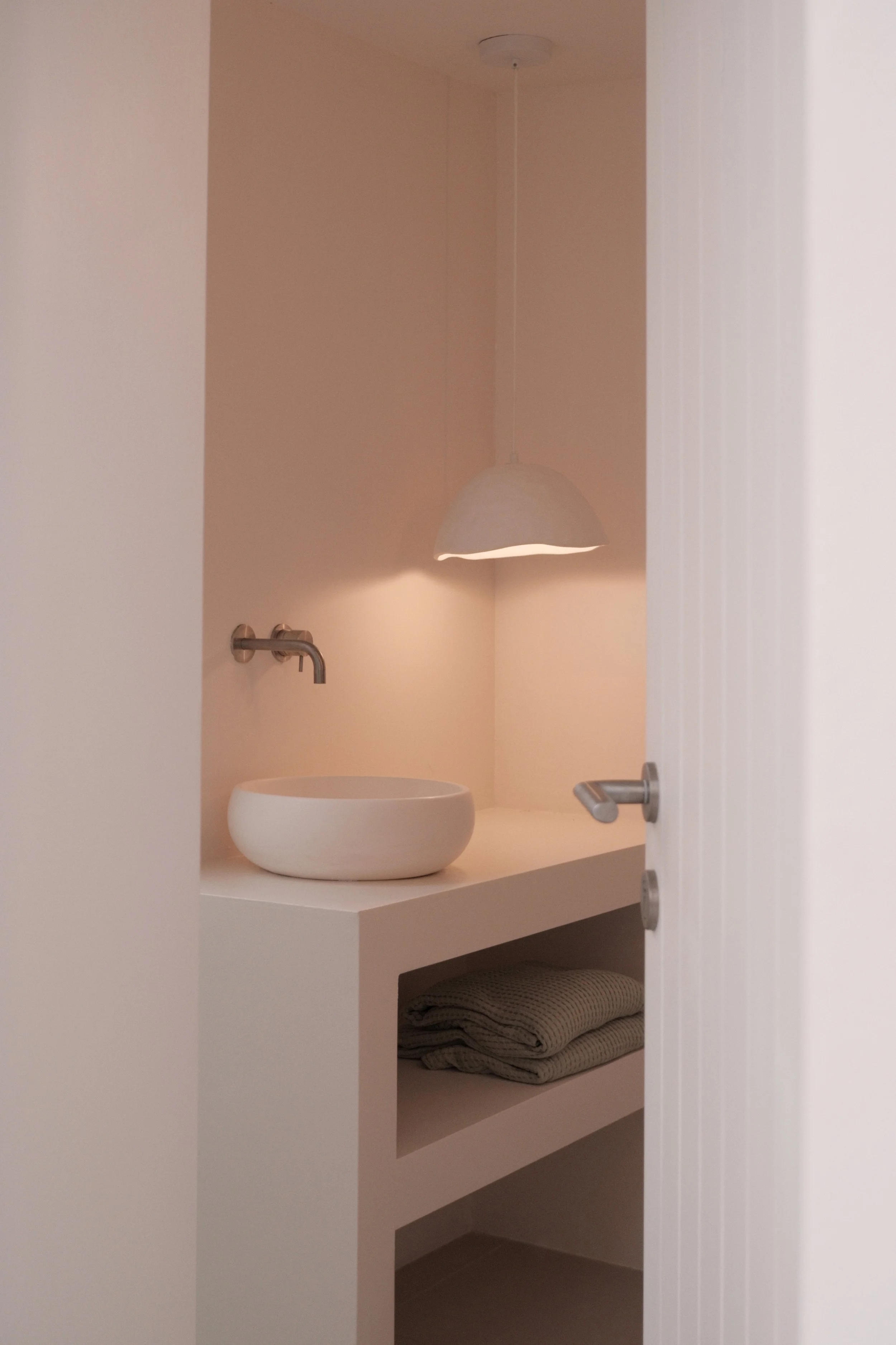A minimalist bathroom with a white vessel sink on a white countertop, a wall-mounted faucet, a hanging lamp above, a towel shelf with folded beige towels underneath, and neutral-colored walls.