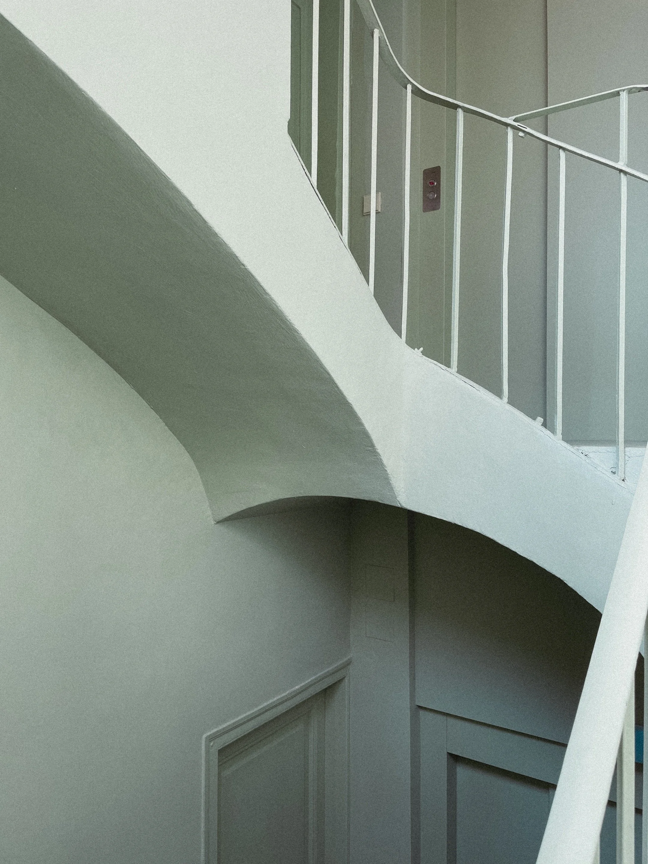 Interior view of a stairwell with a white curved railing, a light green wall, and a door with a panel at the bottom.