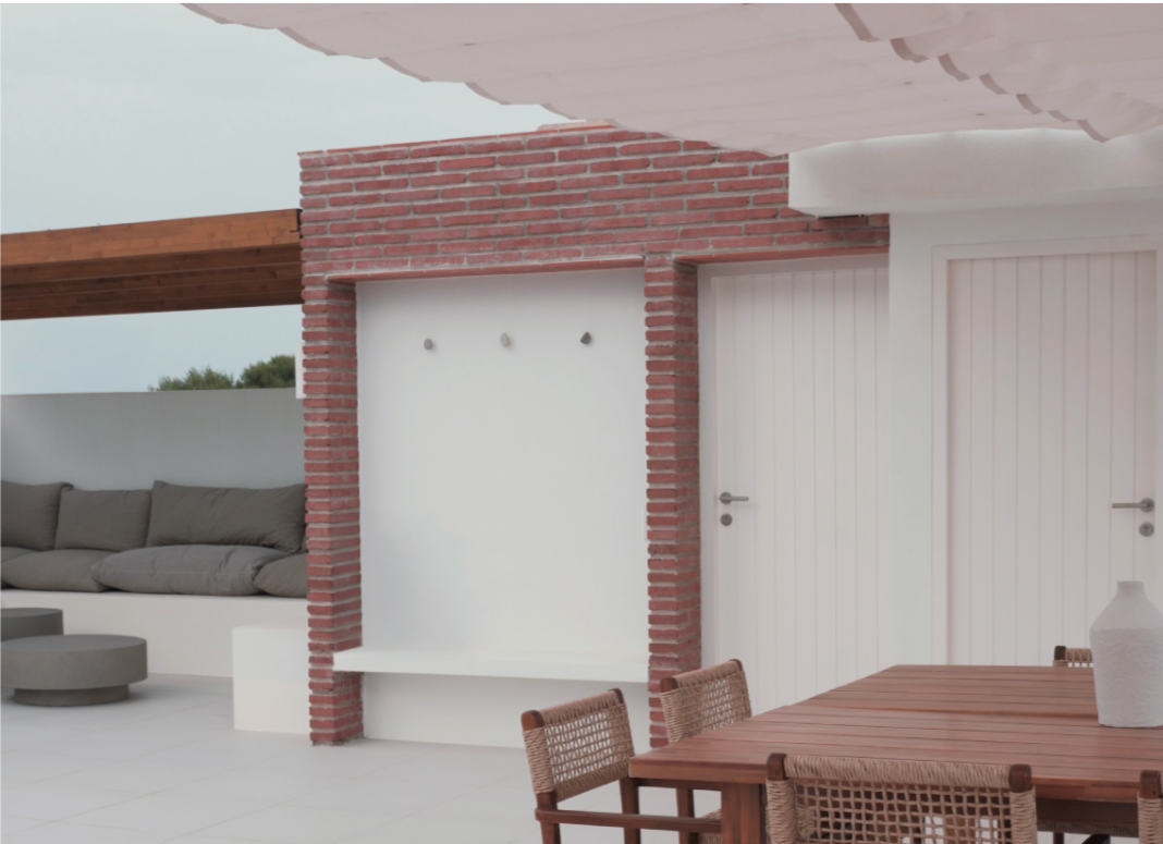 Modern outdoor patio with a brick fireplace, white walls, wooden dining table with wicker chairs, and a gray sofa with cushions.