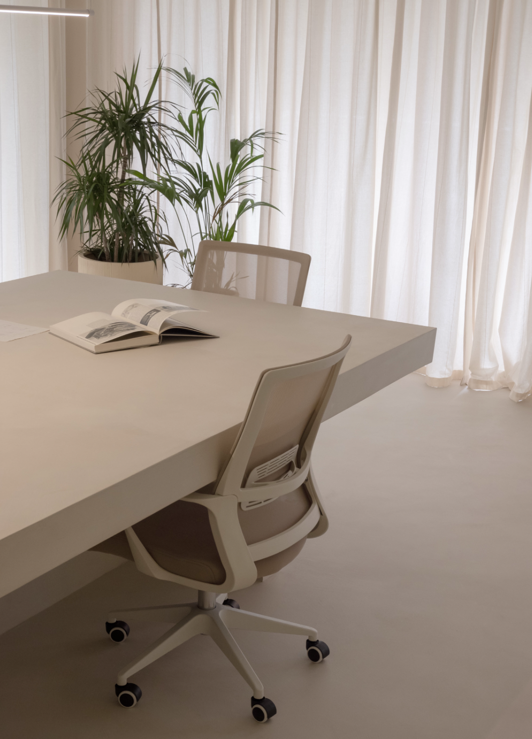 A modern office meeting room with a white table, an open magazine, a beige office chair on wheels, a potted plant with green leaves, and white curtains in the background.