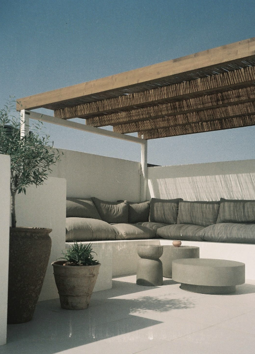 Outdoor seating area with cushioned sofas, potted plants, and round tables on a terrace under a wooden pergola with a blue sky background.