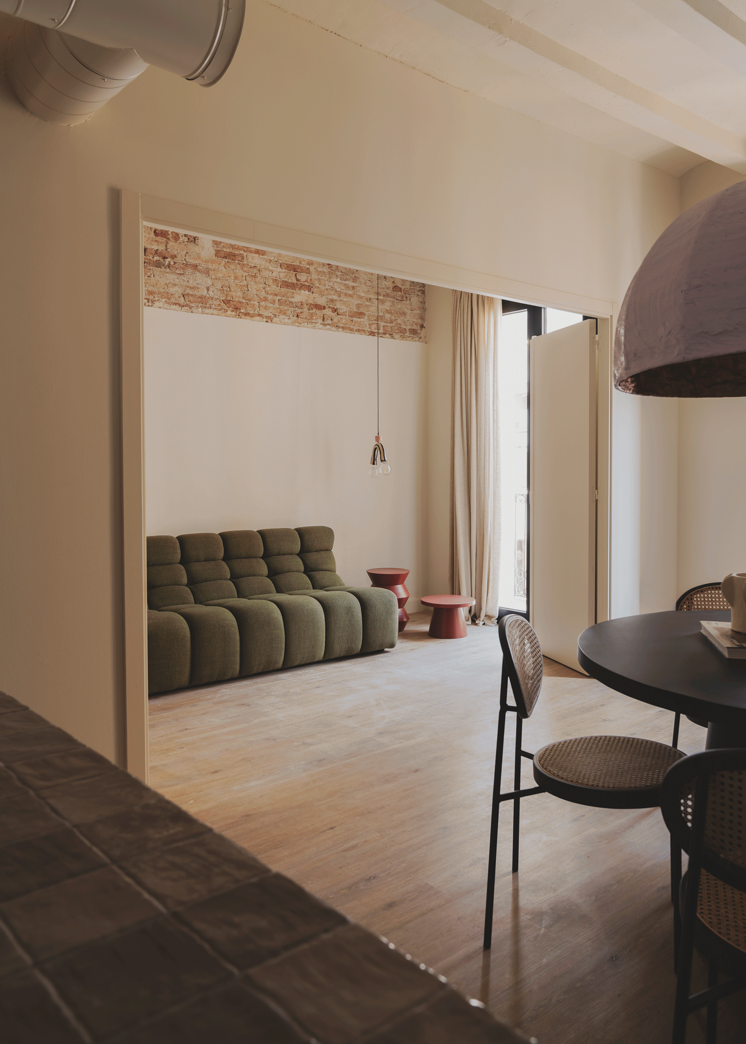 A cozy living room with a green tufted sofa, small red side tables, a hanging light bulb fixture, and a large window with curtains, featuring an exposed brick wall section.
