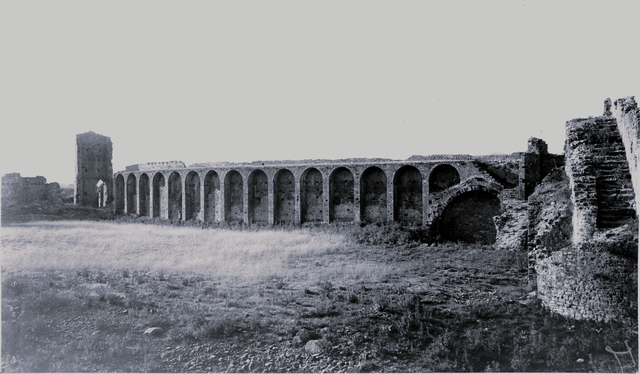 Black and white photo of an ancient Roman aqueduct with multiple arches and a tower, surrounded by open grassy fields.