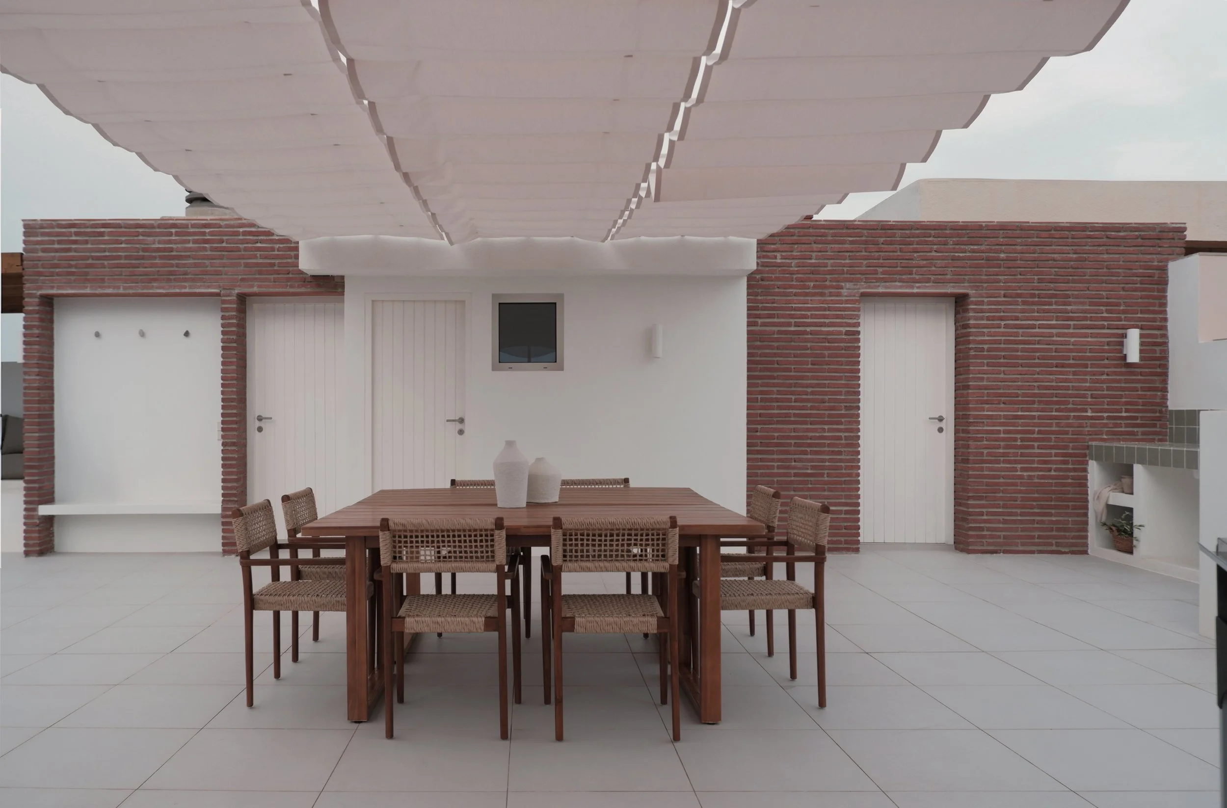 A modern outdoor patio area with a large wooden dining table and multiple rattan chairs, white tiled flooring, brick and white wall backgrounds, and a decorative overhang overhead.