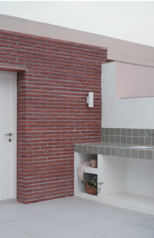 A modern outdoor kitchen area with a red brick wall, a white door, a tiled countertop, and a small built-in shelf with a plant and a basket.