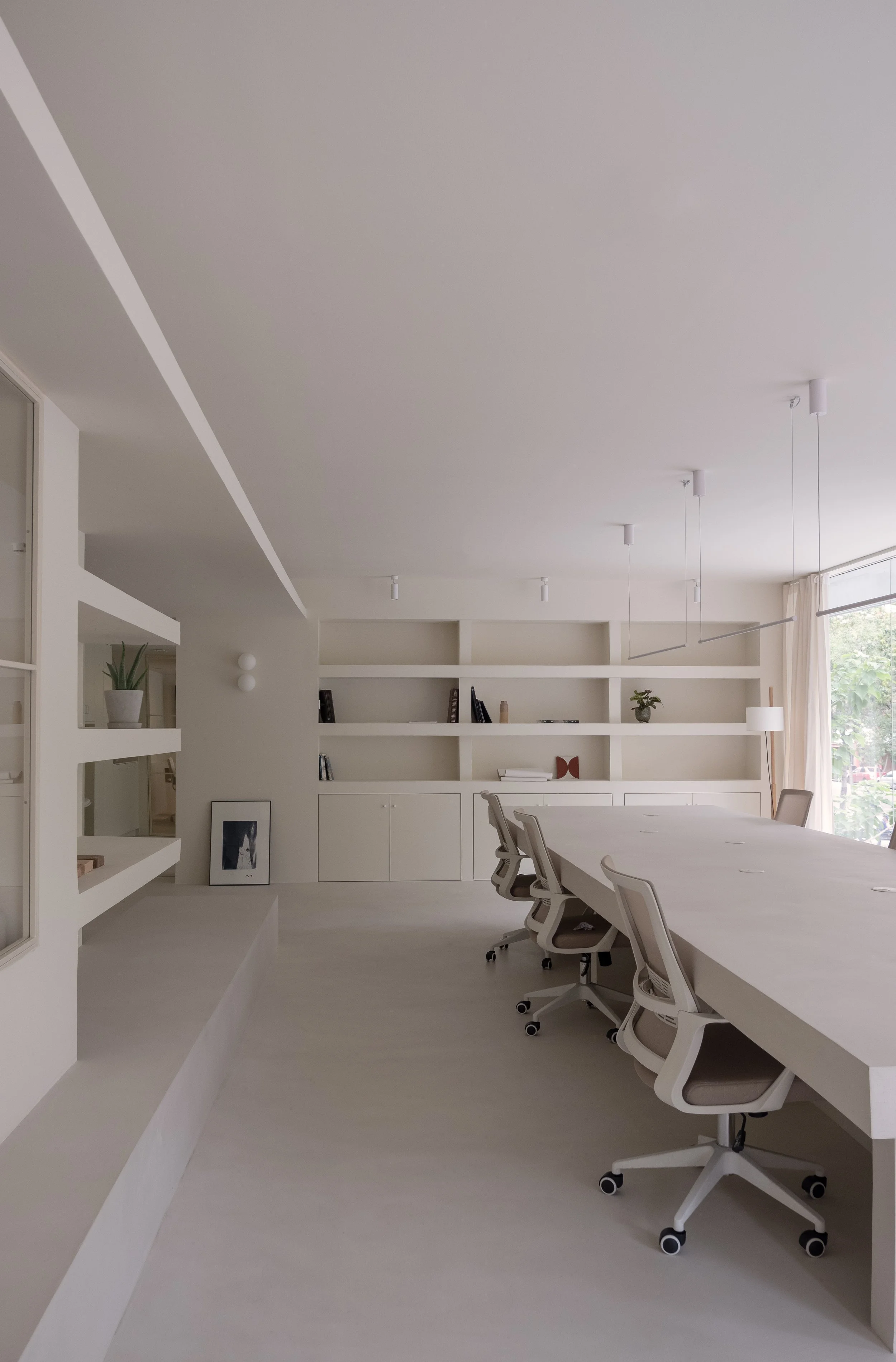 Modern, minimalist conference room with a long white table, white office chairs on wheels, built-in white shelves with decorative items, large window with beige curtains, and pendant lights on the ceiling.