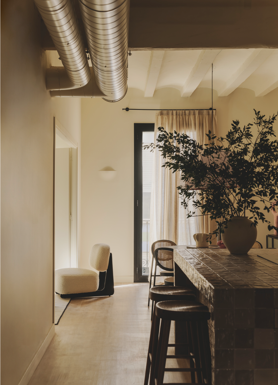 Interior of a room with a beige armchair, a large leafy plant in a vase, a table with bar stools, a window with beige curtains, and exposed ceiling pipes.