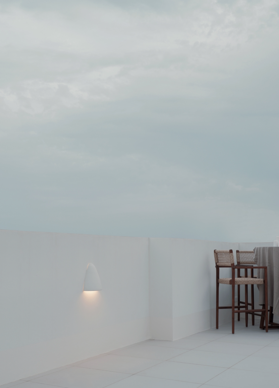 Rooftop terrace with a white wall, outdoor lamp, and two bar stools with woven seats under a cloudy sky.