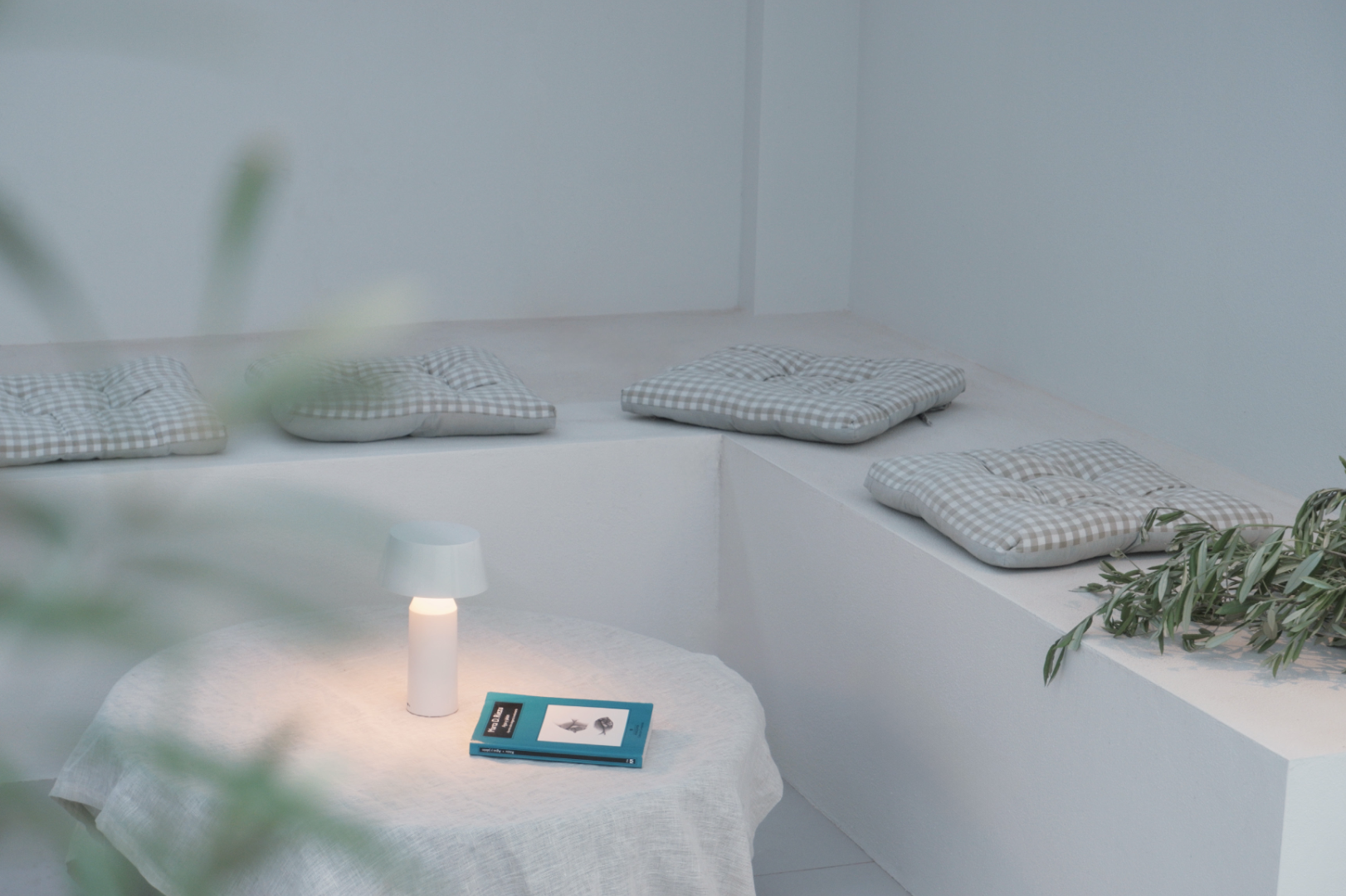 A minimalist seating area with a built-in white platform with four checkered cushions, a small white table with a lamp, a couple of books, and some greenery on the edge.