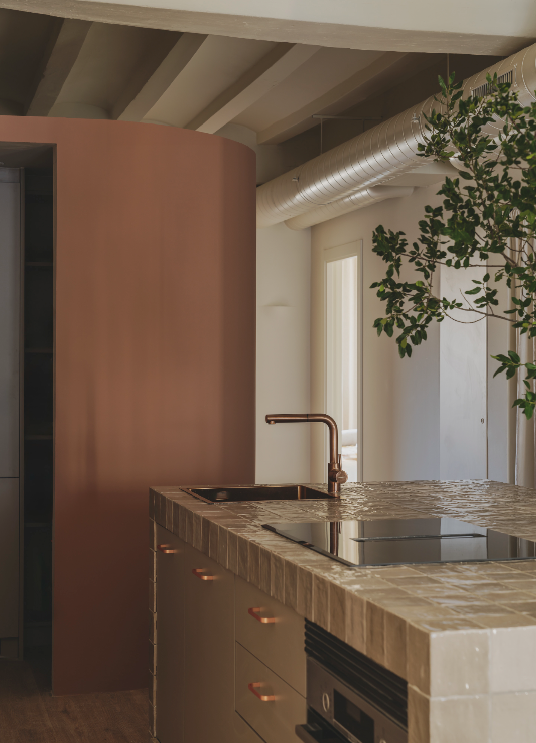 Modern kitchen with a beige tiled island counter, copper faucet, black cooktop, white oven, soft pink curved wall, exposed white ceiling beams, a large vent pipe, a window, and a green plant.