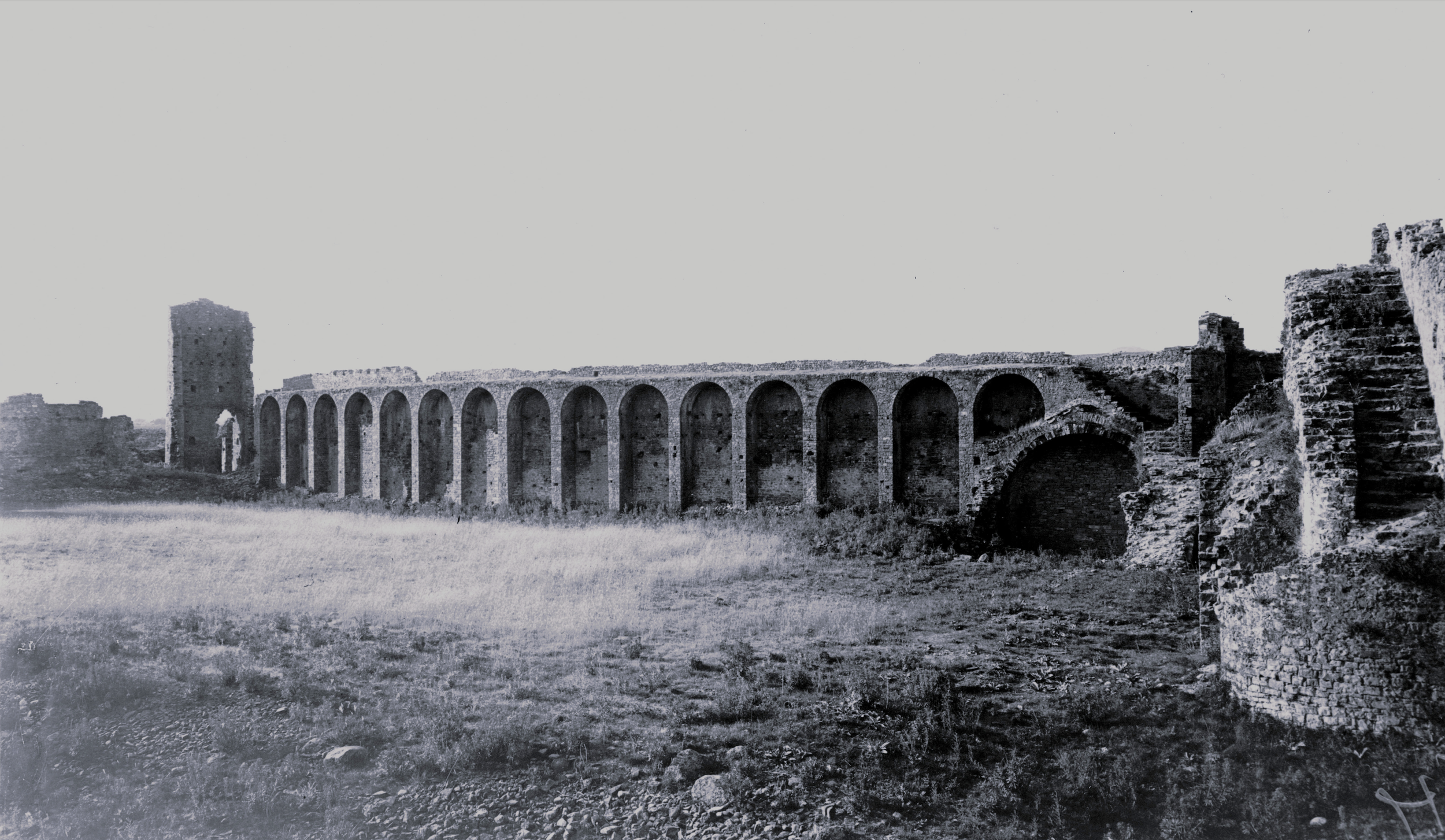 Old stone aqueduct ruins with multiple arches extending across a grassy field, with some deteriorated sections and a tall tower on the left.