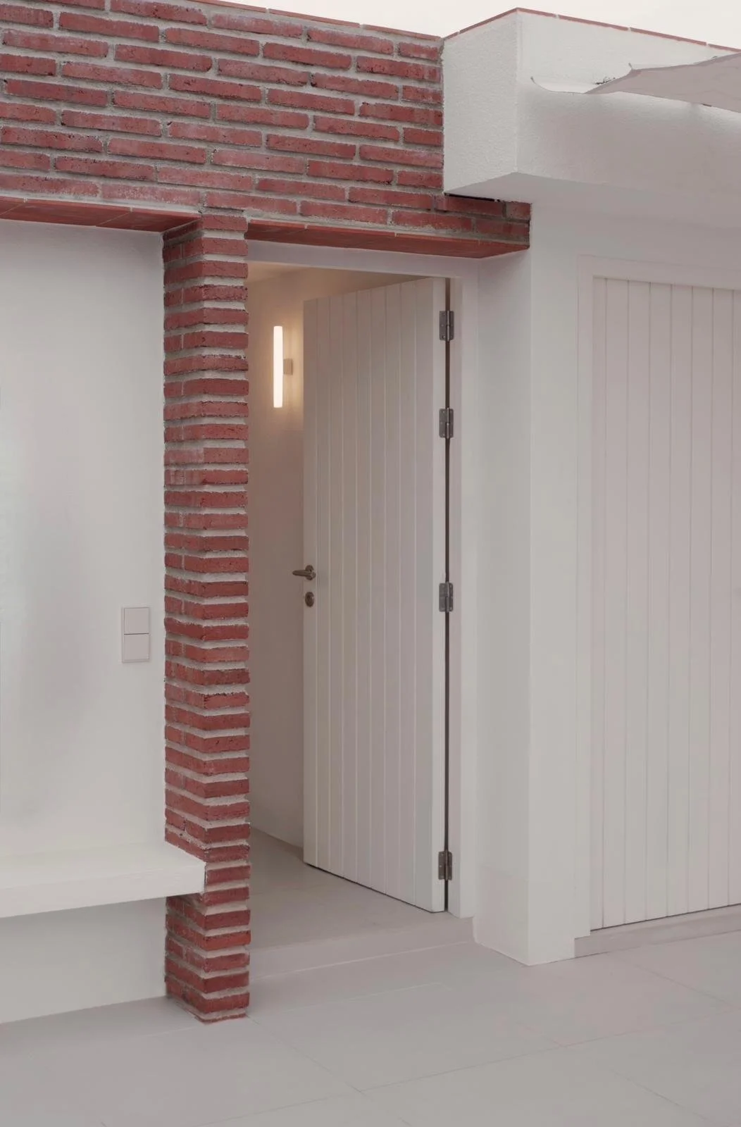 Interior view of a modern room with a white wooden door, a white wall, a red brick column, and a wall-mounted light fixture.