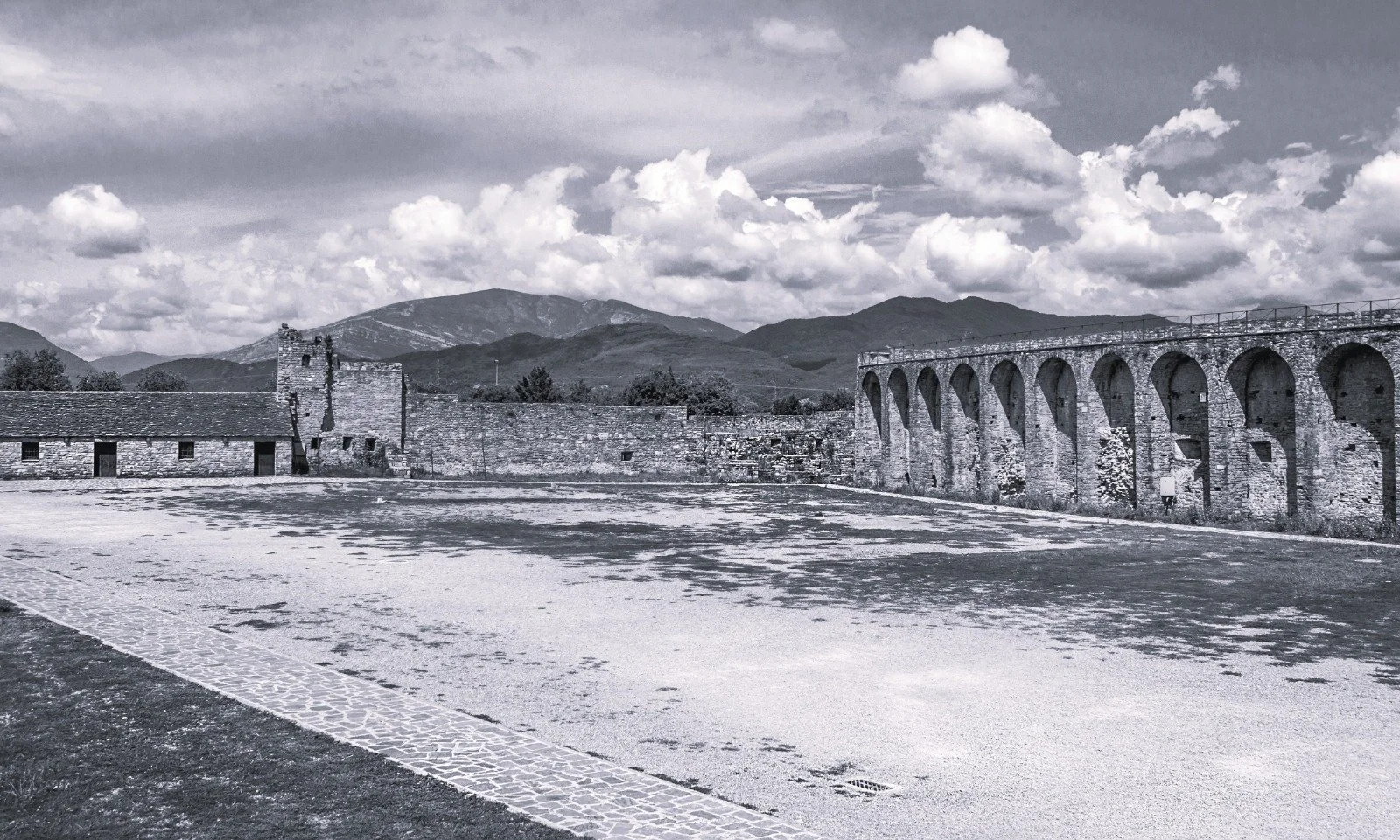 Black and white photo of an ancient stone aqueduct with multiple arches, a small stone building, and a mountain range with cloudy sky in the background.